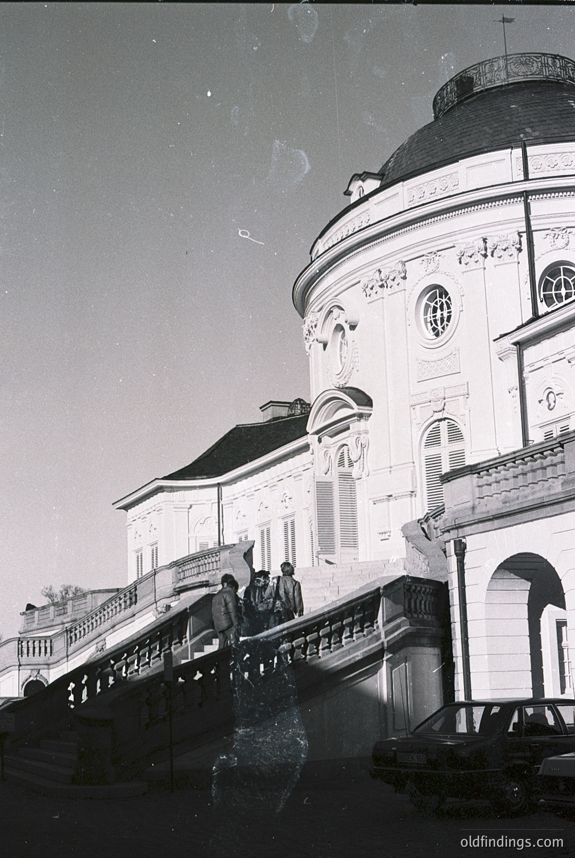 Neoclassical building corner with ornate dome and balustrade, likely 19th-century European architecture. Group of people ascending a curved stone staircase. Aged black-and-white photo suggests historical or vintage appeal.