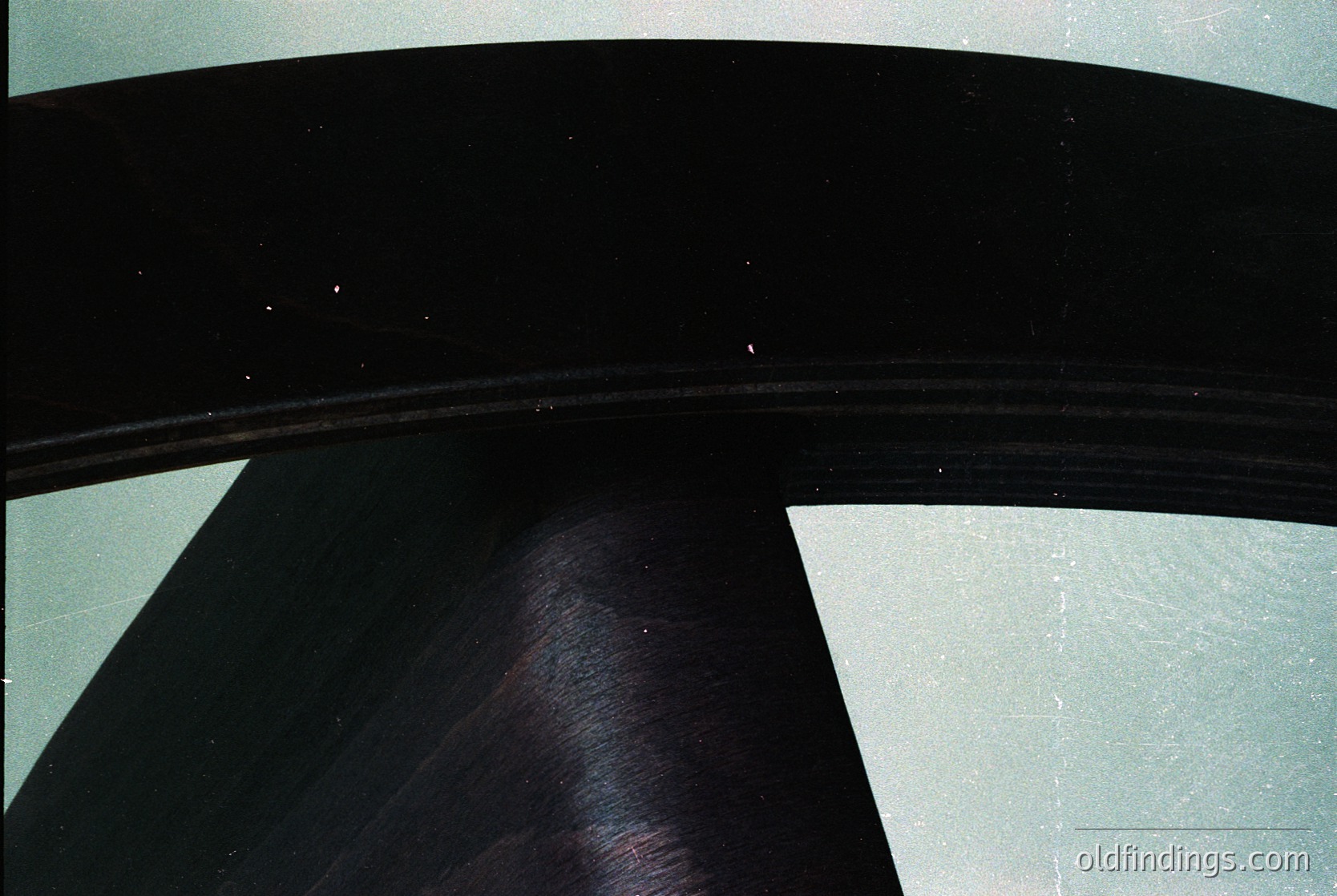 Close-up of a curved, black metal or plastic surface with a smooth, slightly reflective finish. The edge shows a sharp, clean bend, likely part of an industrial or architectural structure. The muted lighting suggests an interior setting, possibly from the 1980s–1990s.