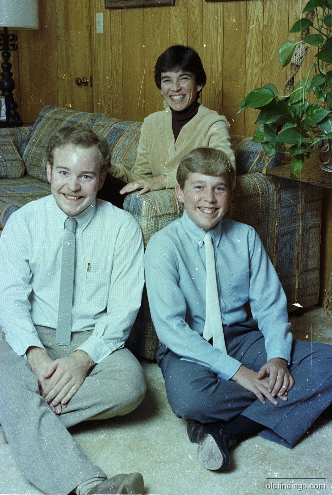 Family portrait from the 1970s featuring a woman seated on a brown leather couch with two young boys kneeling beside her. Woman wears a cream-colored cardigan over a patterned blouse; boys in light blue dress shirts with ties. Indoor setting with wooden paneling, potted plant, and neutral-toned carpet.