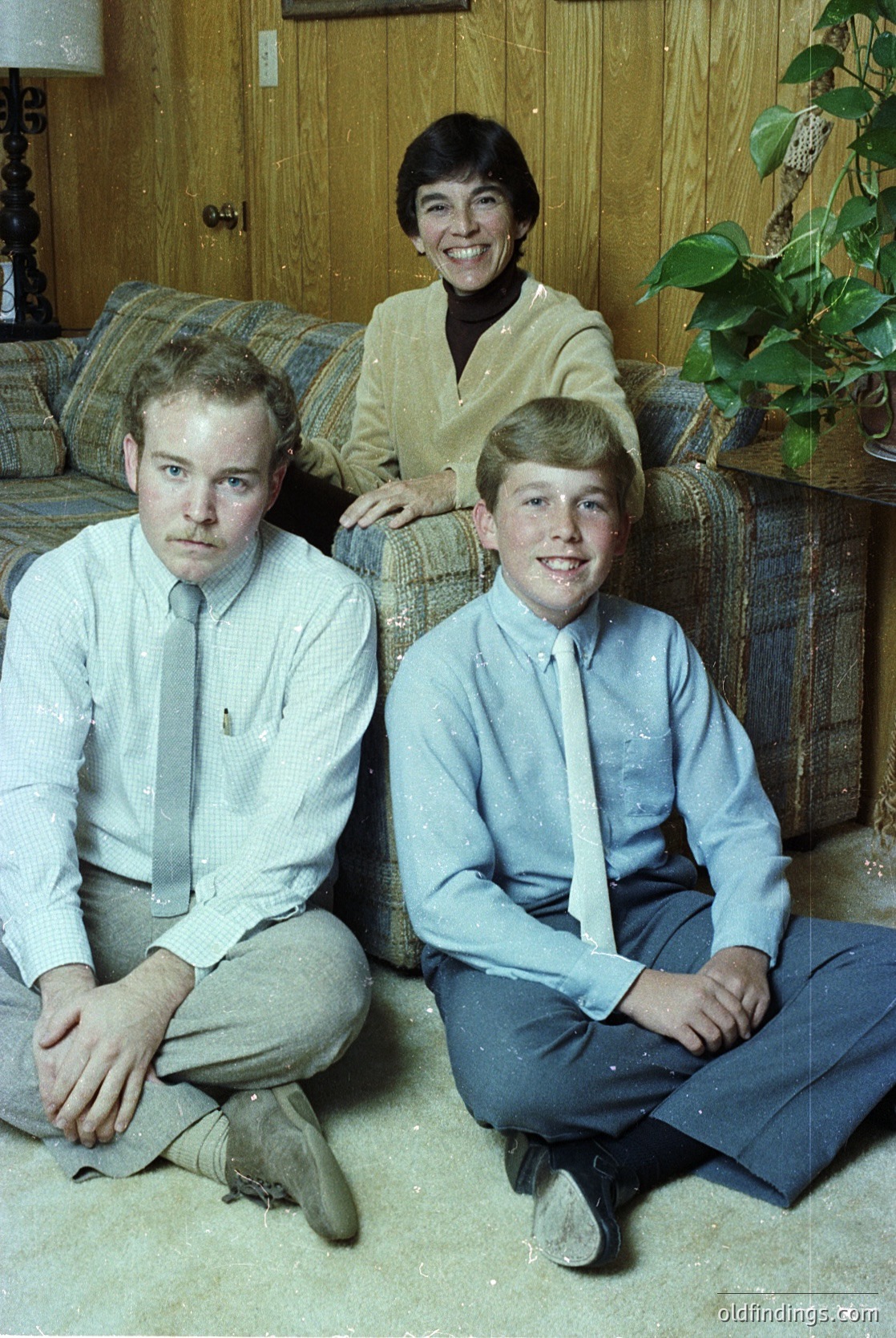 Family portrait from the 1970s featuring a woman in a plaid skirt and cream sweater, flanked by two young men in dress shirts and ties. Indoor setting with wooden paneling, a potted plant, and a plaid sofa. Warm, candid expressions capture a nostalgic, mid-century American home vibe.