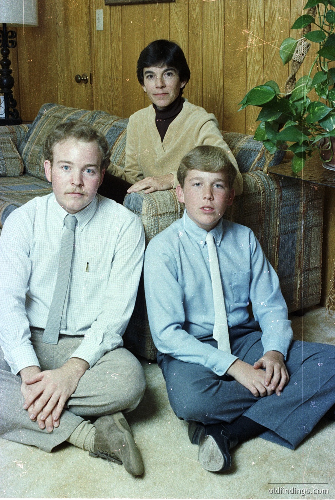 Family portrait from the 1970s featuring a man in a light gray suit and tie, a woman in a plaid skirt and cream cardigan, and a boy in a blue dress shirt and tie. Indoor setting with wooden paneling, a potted plant, and a plaid sofa.