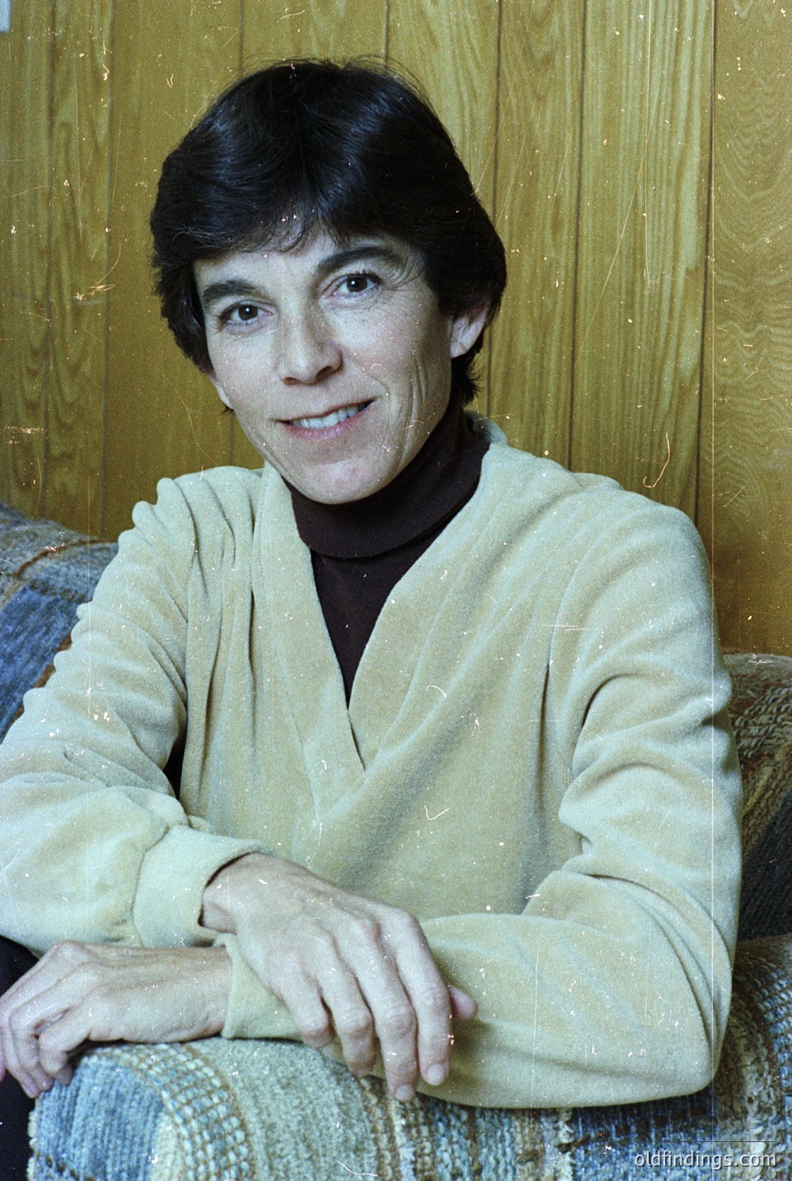Portrait of a person in a relaxed pose, seated on a woven textile with wooden paneling backdrop. Light-colored, textured sweater with a V-neck and dark top layer. Mid-length, dark hair styled neatly. Warm indoor lighting suggests a 1970s–1980s aesthetic.