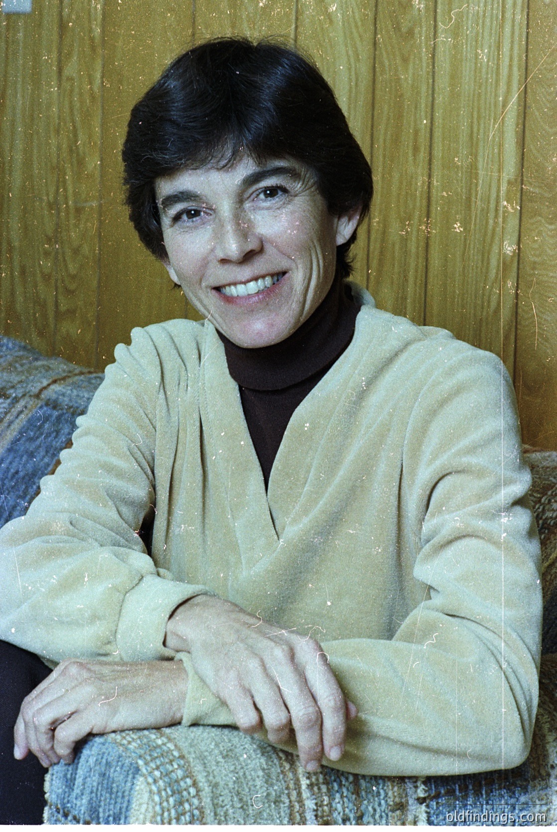 Portrait of a woman in a light-colored, textured sweater, seated indoors against a wooden-paneled wall. Her short, dark hair and warm smile suggest a candid, mid-20th-century studio or home setting.