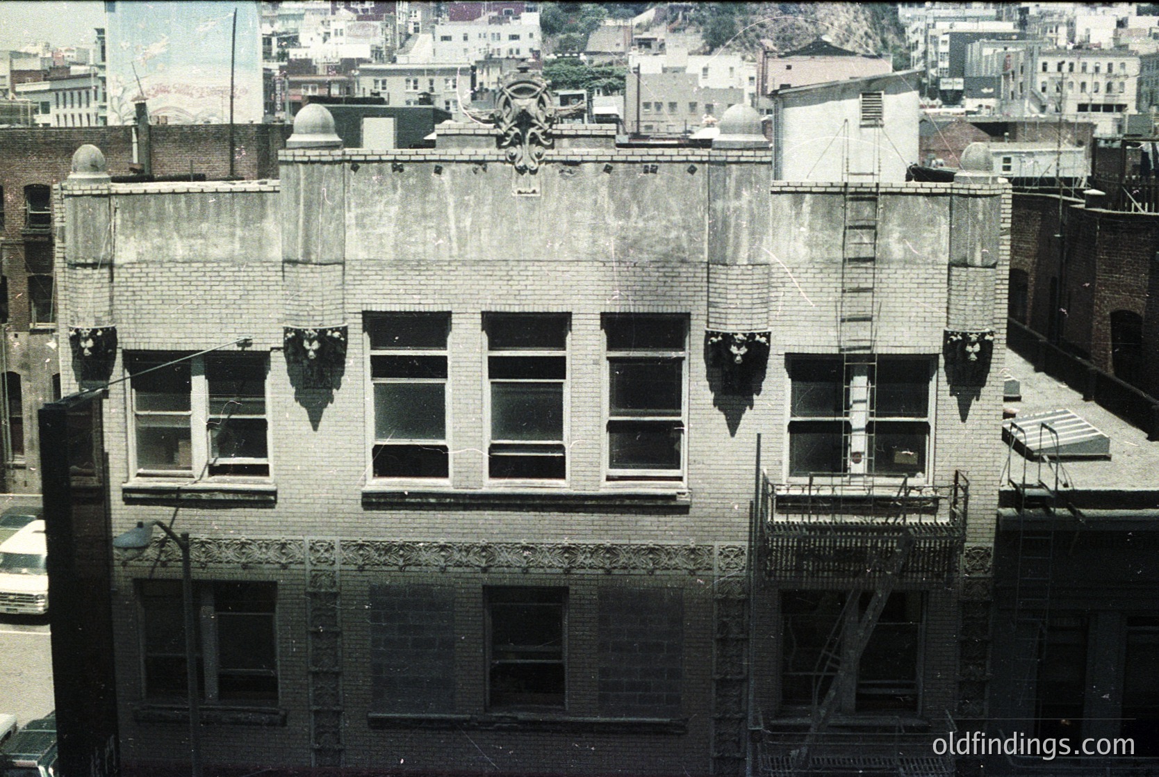 Three-story brick commercial building façade, likely early 20th century. Symmetrical design with decorative cornice and fire escape. Urban setting with adjacent structures and distant hills. Potential or architecture.