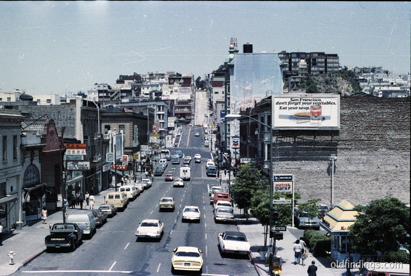 Vintage urban street scene with mid-century architecture, featuring a wide boulevard lined with vintage cars and multi-story buildings. Prominent billboard advertises "Don’t forget your vegetables" for "Ken Franks." Coastal backdrop with water and distant hills.
