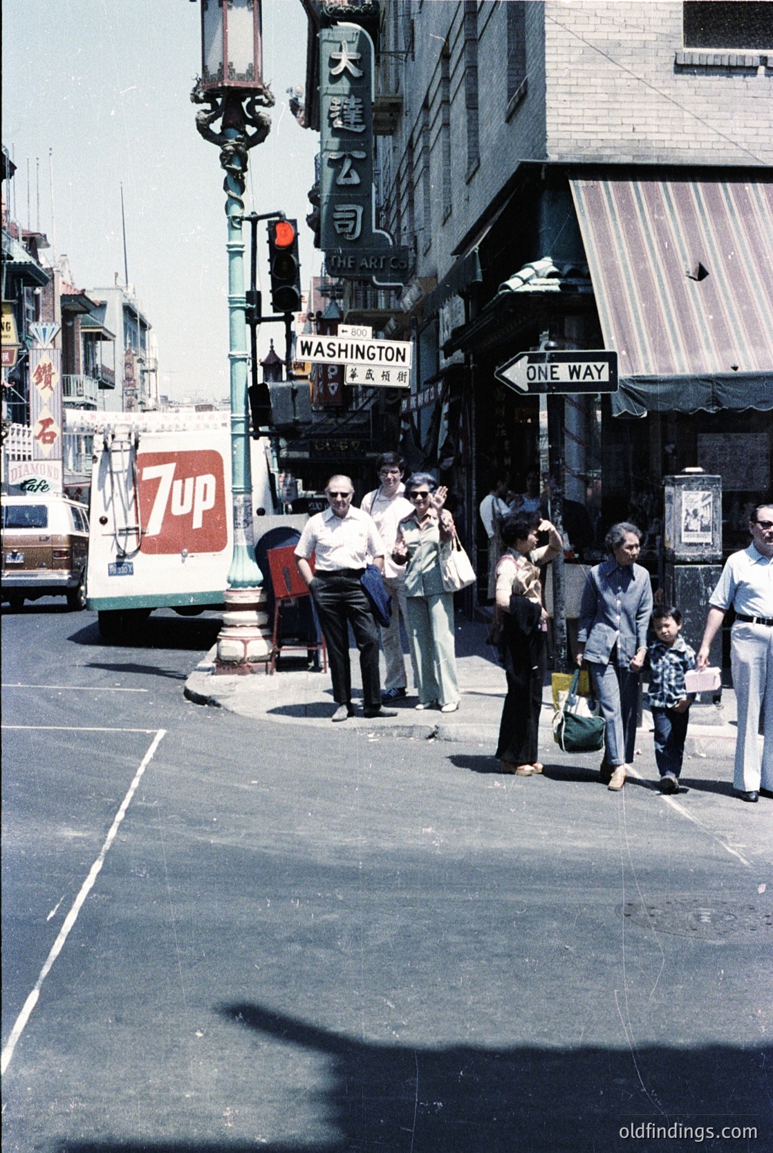 Vintage street scene at Washington Ave intersection, likely San Francisco’s Chinatown. Crowd of 1970s-era pedestrians—men in suits, women in dresses, a child—waiting at a crosswalk. Neon "7UP" sign and Chinese characters on a building sign. "One Way" street sign visible. Urban architecture with storefronts and parked vehicles.