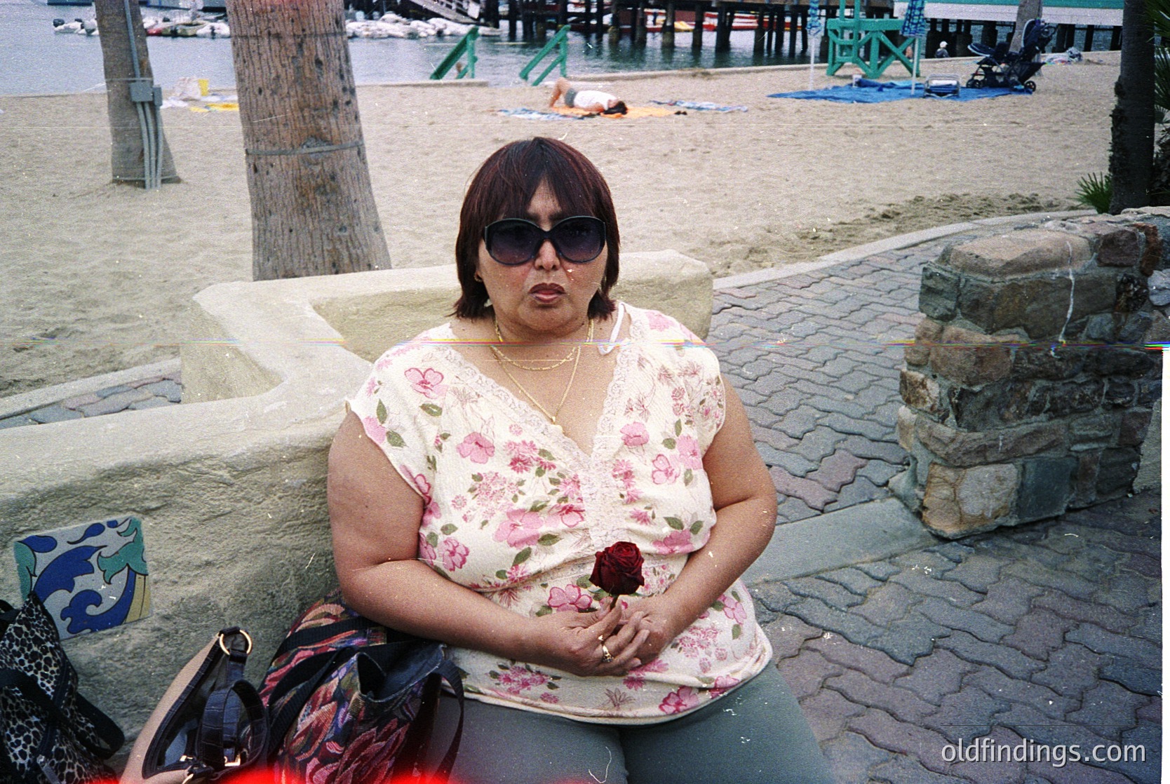 A woman in a floral blouse and sunglasses sits on a stone bench beside a seaside promenade, holding a red rose. Beachgoers relax on towels in the background, with a pier and boats visible across the water. The setting suggests a mid-20th-century seaside resort.