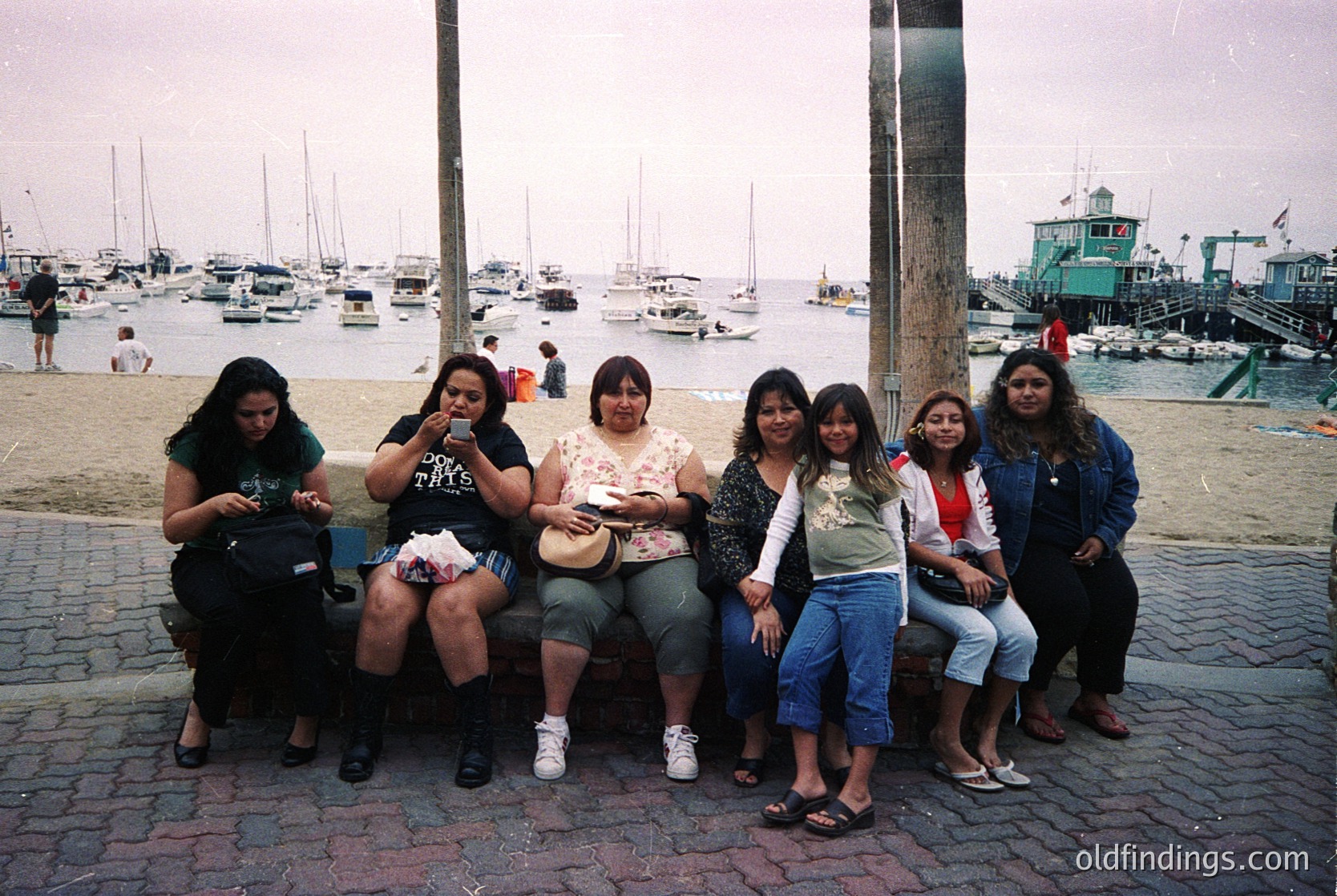 Group of six women posing on a seaside promenade, 1990s. Cobblestone path, marina with sailboats, and a lighthouse in background. Casual attire: jeans, graphic tees, and sneakers.