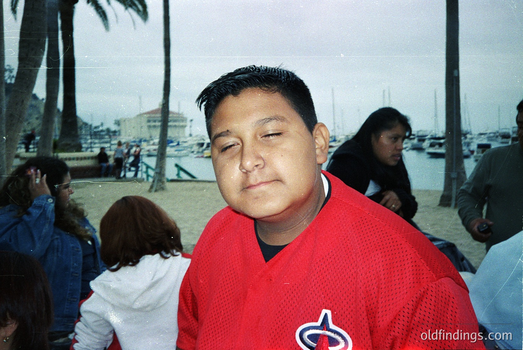 Portrait of a man in a red baseball jersey (likely 1990s-2000s era) with a logo resembling the **Seattle Mariners**, standing outdoors near a marina with palm trees and boats. Crowd and waterfront setting suggests a casual, public event.