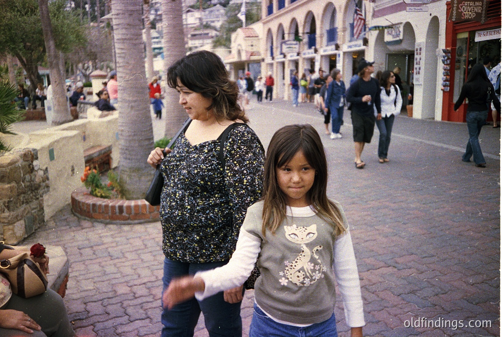 A woman and child stroll through a Mediterranean-style plaza with arched storefronts and palm trees. The woman wears a black polka-dot top, the child a grey sweatshirt with a cartoon character. Background shows pedestrians, potted plants, and a mix of modern/retail architecture. Likely a tourist district, possibly California’s Spanish Colonial Revival style.