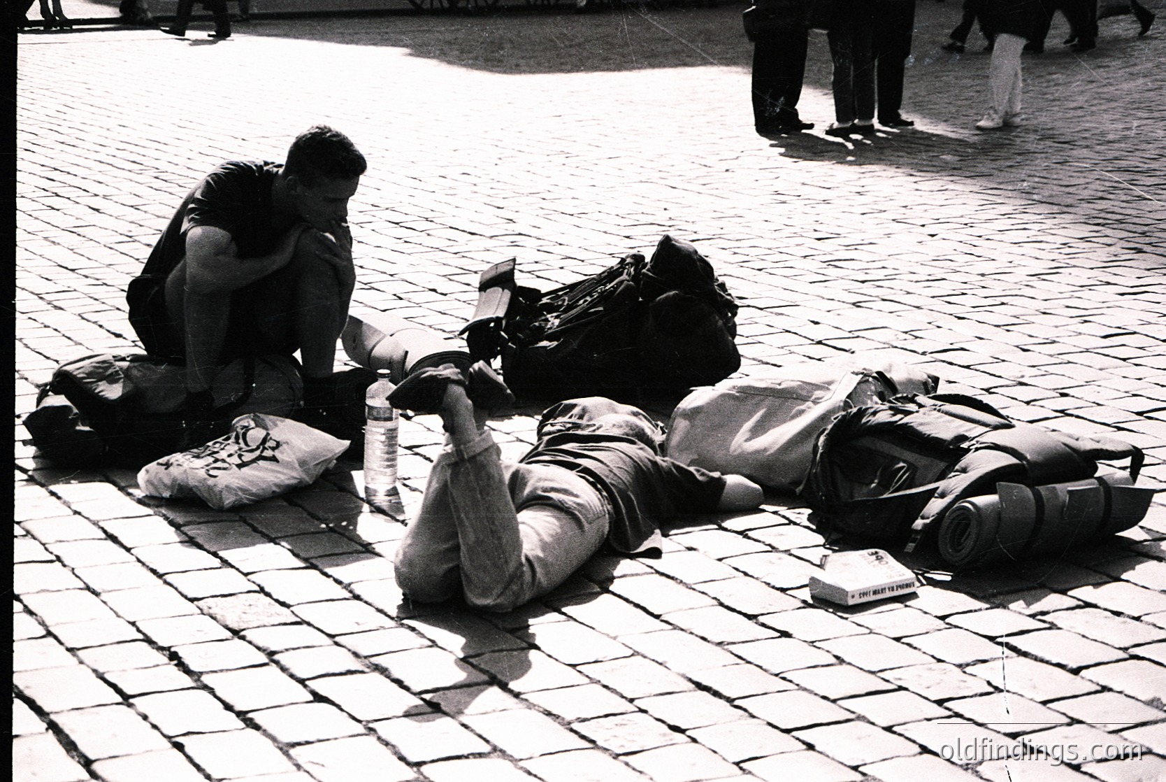 Two individuals rest on cobblestone pavement, surrounded by backpacks and personal items. The person on the left kneels, leaning forward with arms around knees, while the prone person lies on their back, legs extended. A pillow and book lie nearby. Background shows blurred pedestrians in a public square, suggesting a casual, urban setting.