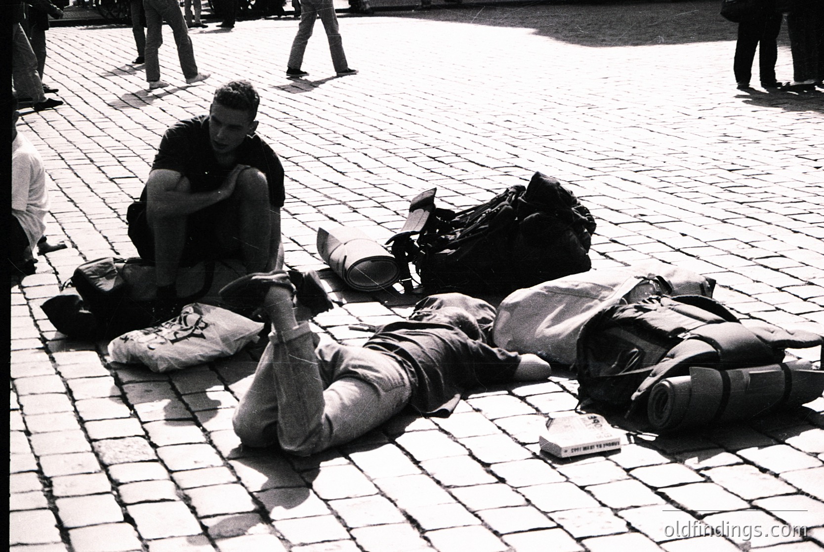 Two travelers rest on cobblestone pavement, surrounded by backpacks and luggage. One sits cross-legged, the other reclines. Mid-20th century street scene with urban architecture in background.