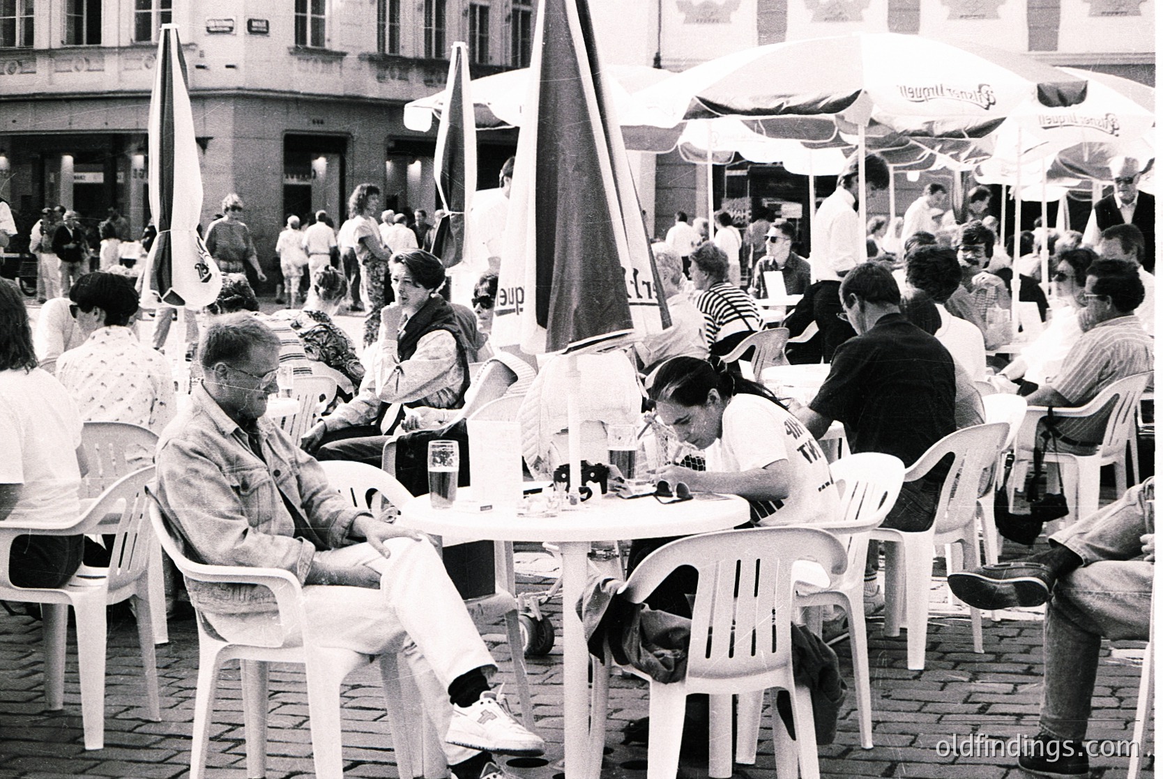 Vintage black-and-white street café scene with plastic chairs and tables under striped umbrellas. Mid-20th-century European urban setting, likely 1960s-70s. Crowded plaza with people dining, drinking, and socializing. Architectural backdrop of multi-story buildings with balconies. Ideal for historical research or nostalgic design references. é