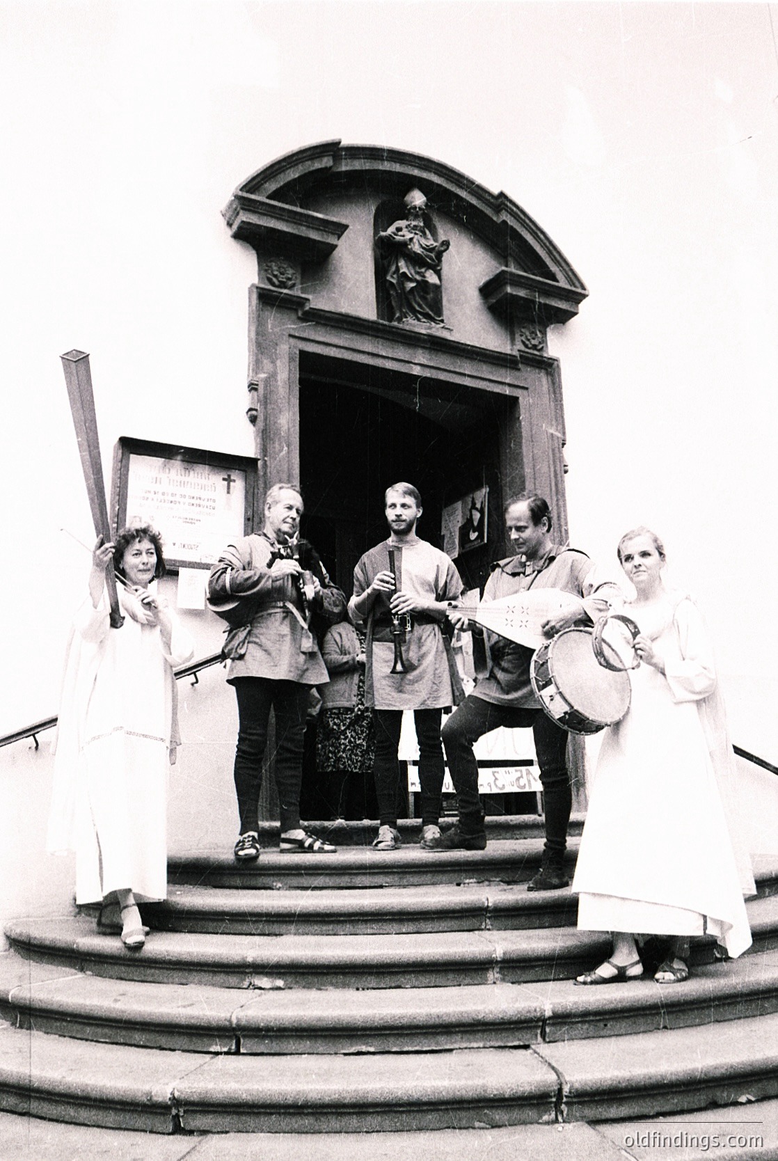 Vintage black-and-white photo of a folk ensemble performing at a church entrance, likely Eastern European . Group includes musicians playing traditional instruments (accordion, drum, flute) and a banner with Cyrillic script. Architectural details: arched doorway with decorative stonework and statue above.