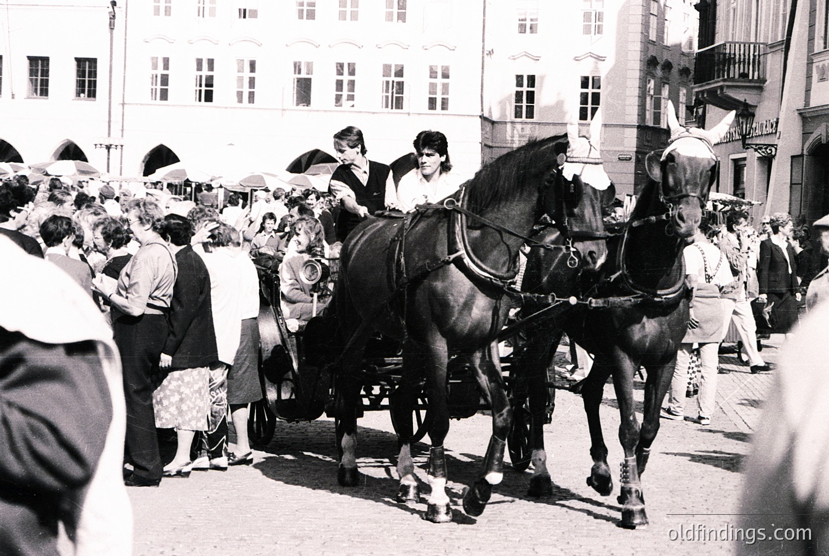 Horse-drawn carriage with two black horses in a bustling European street, mid-20th century. Crowd in vintage attire, multi-story buildings with balconies and awnings. Likely a festival or public event.