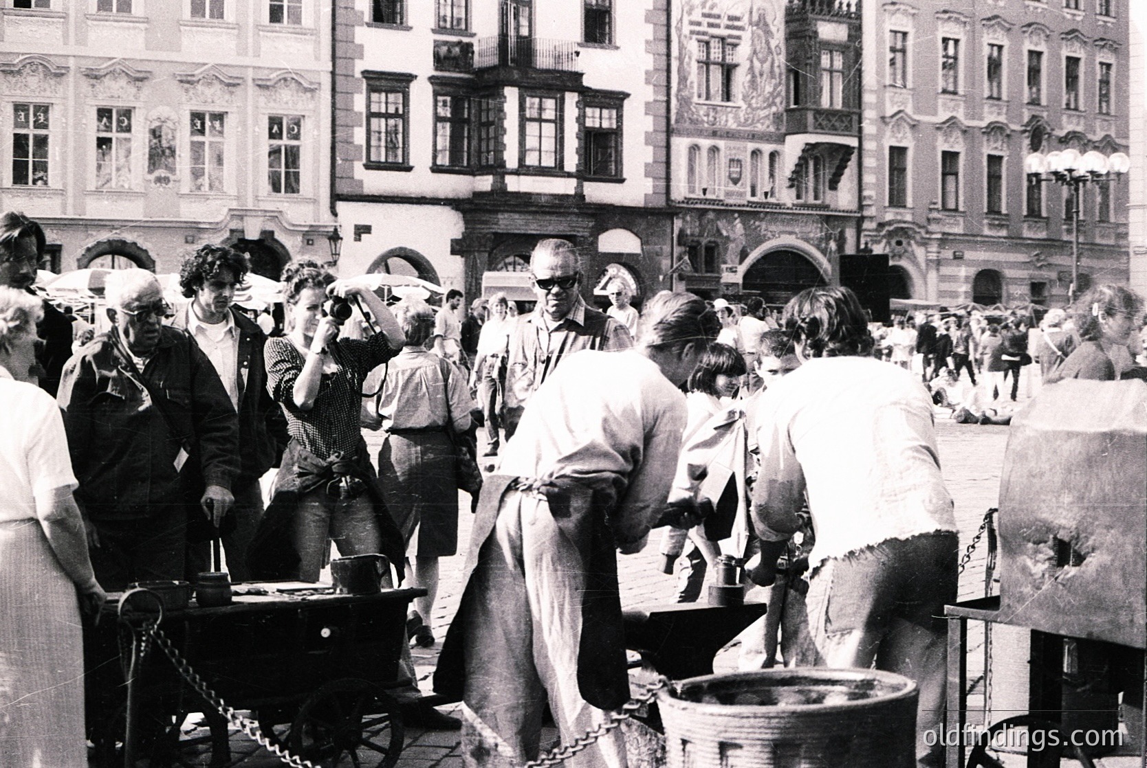 Black-and-white street scene featuring a lively outdoor market in a European city square, likely 1960s–1970s. Crowd gathers around a vendor cooking food in a large pot over an open flame, with ornate historic buildings in the background. Distinctive Art Nouveau facades and cobblestone pavement enhance the scene.