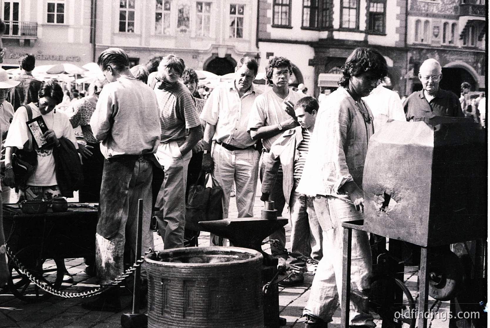 Crowded outdoor street market scene featuring blacksmithing demo—artisans work with an anvil and bellows, surrounded by onlookers in casual 1970s attire. Historic European architecture with arched windows and balconies in background.