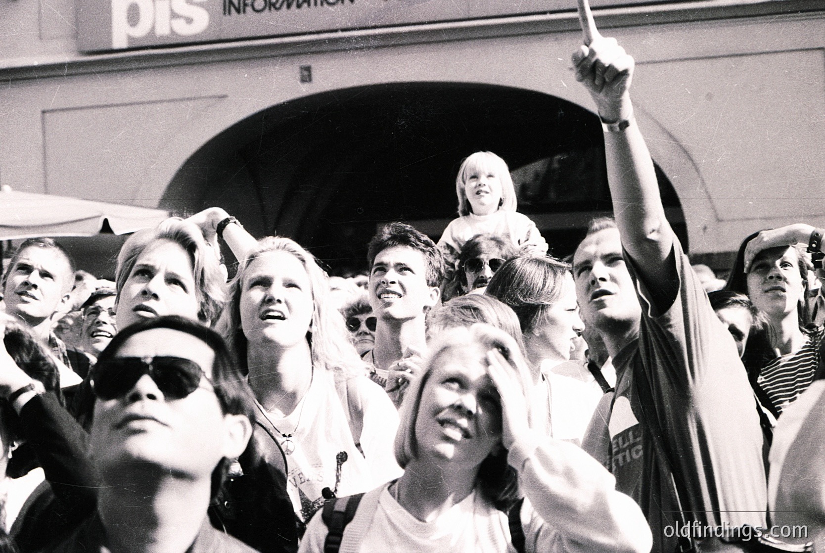 A lively crowd of young people gathers outdoors, cheering at an event. Faces show excitement, with one person raising a flag or banner. Mid-20th century fashion—short hair, sunglasses, and casual attire—suggests a 1960s–1970s setting. A "PIS Information" sign in the background hints at a public or institutional venue. [Crowd cheering at public event, 1960s–1970s fashion, mid-century outdoor gathering, youth culture, flag waving]