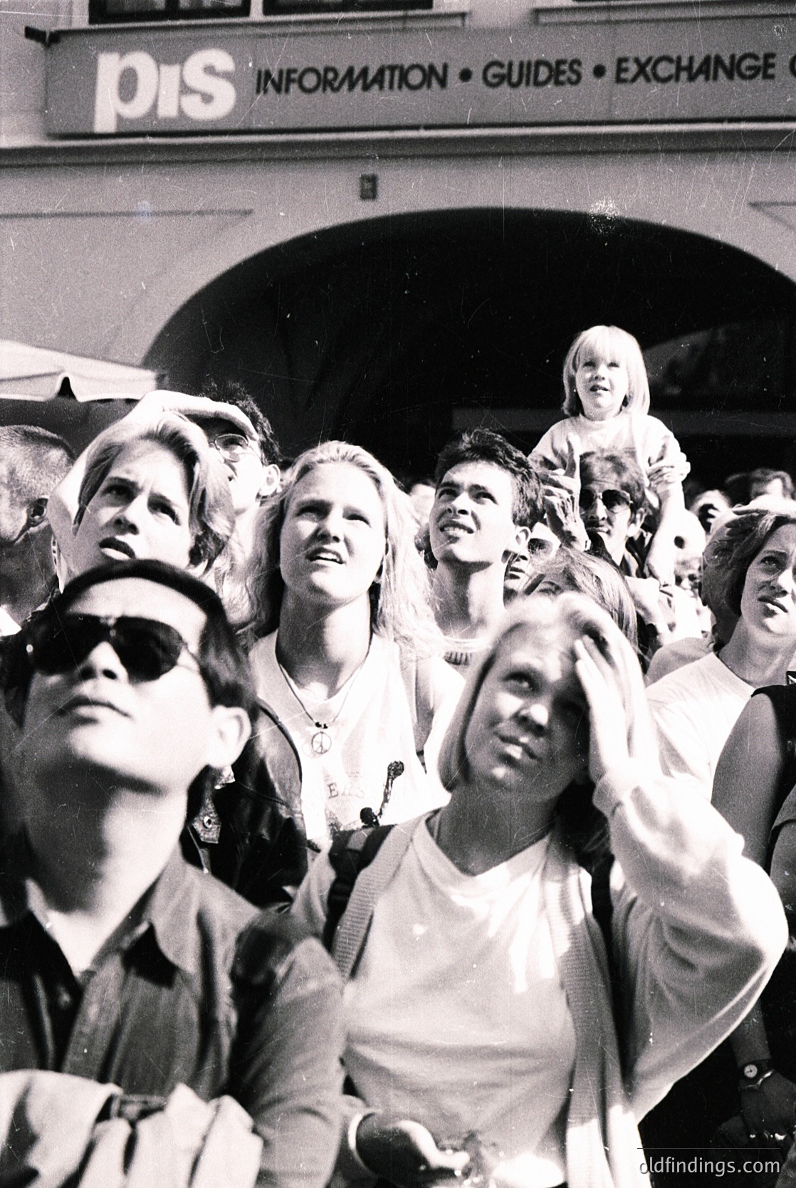 A lively 1960s crowd gathers outside a "PIS Information" building, likely a Polish tourist office. People in casual 60s fashion—sunglasses, short hair, and light clothing—stand close together, some shielding eyes or smiling. The signage suggests travel-related activity, possibly Warsaw or a major European city.