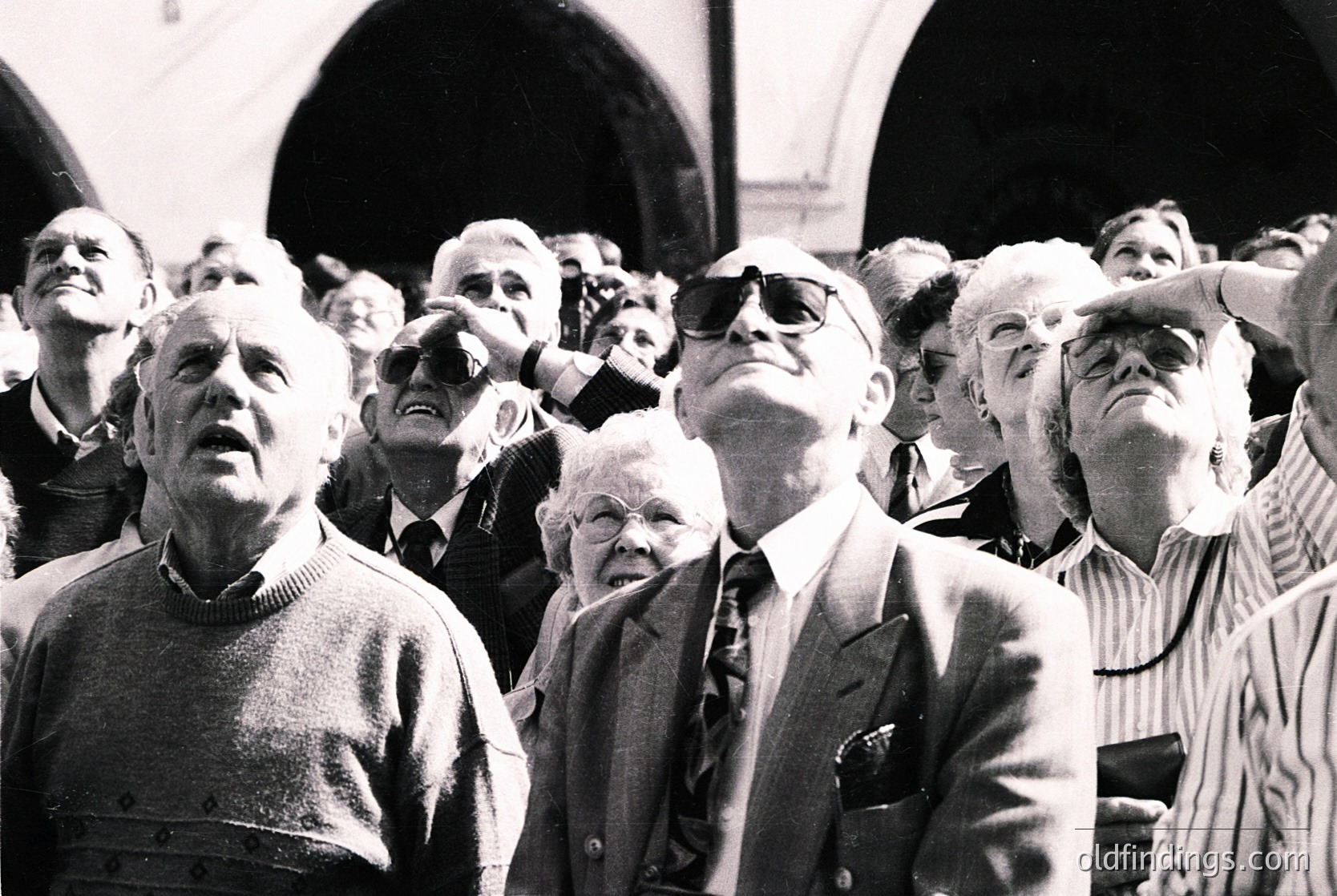 Crowd of elderly men in 1960s-70s attire, gathered indoors under arched stone ceilings, gazing upward with focused expressions. Formal wear includes suits, sweaters, and hats, suggesting a formal event or ceremony. Architectural details hint at a public or institutional setting.