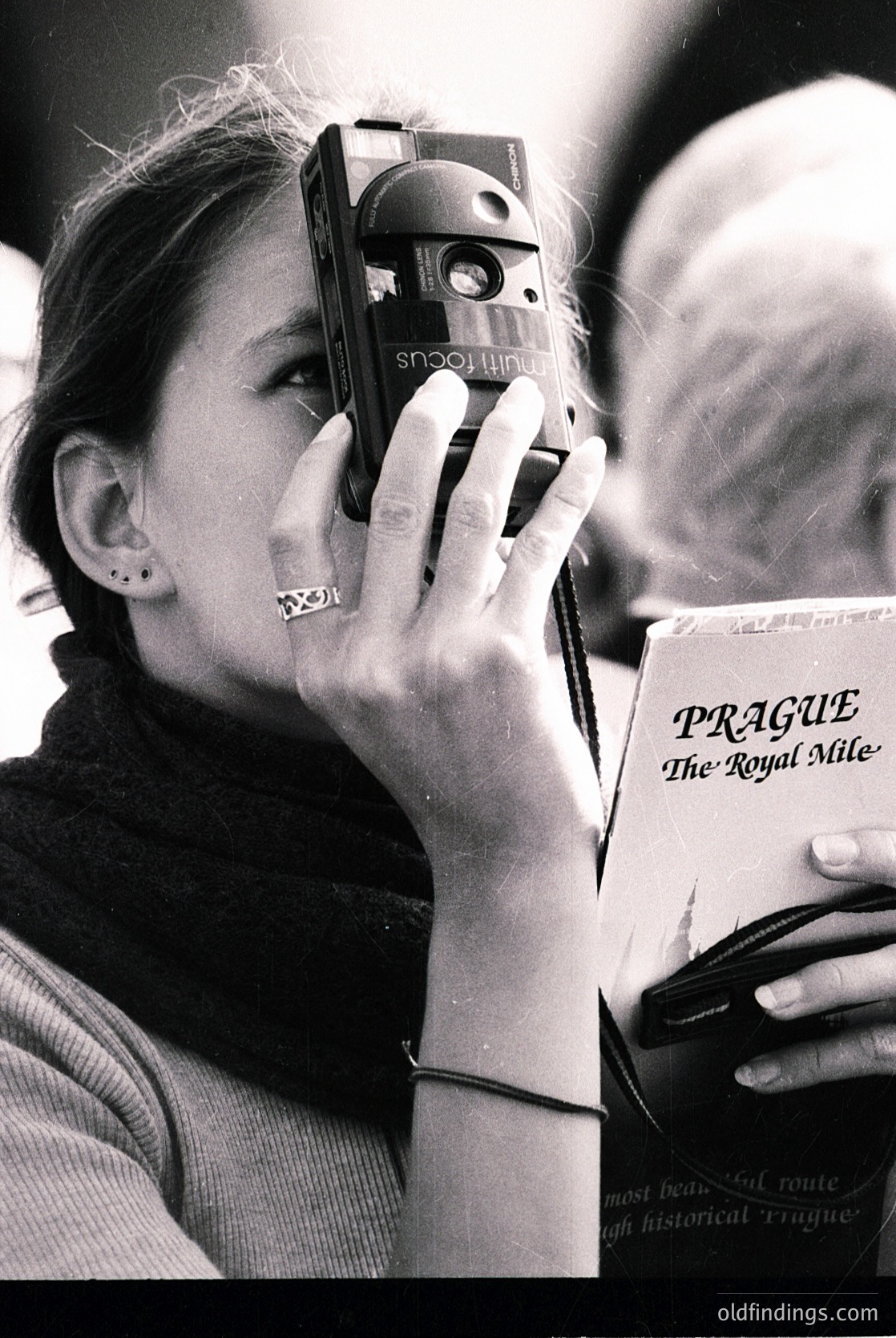 Vintage black-and-white photo of a woman holding a **Sony Mavica** camera (1980s model) while reading *"Prague: The Royal Mile"* guidebook. Mid-century fashion with layered neckwear and minimalist jewelry. Likely 1980s Europe, capturing traveler’s curiosity.
