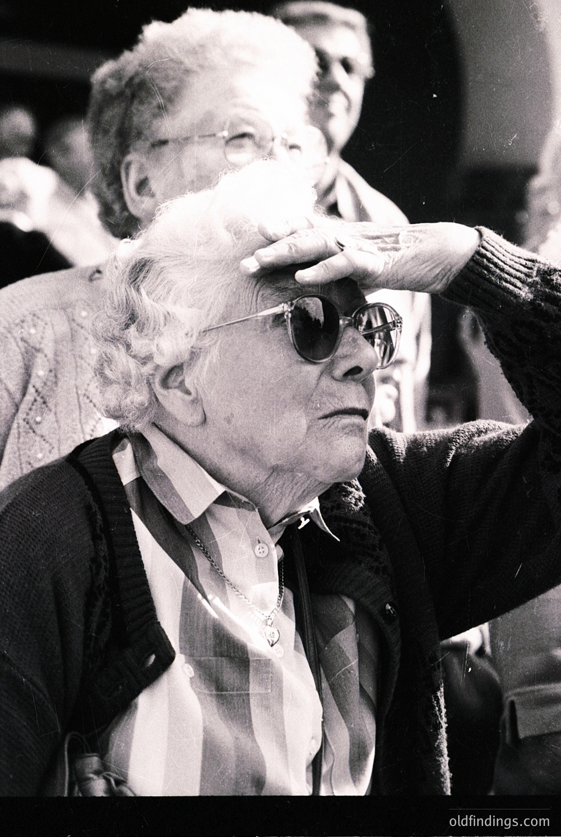 Tight close-up of an elderly woman in mid-20th century attire, wearing round sunglasses and a checkered jacket, with a hand adjusting her hair. Crowded indoor setting with blurred spectators in the background.