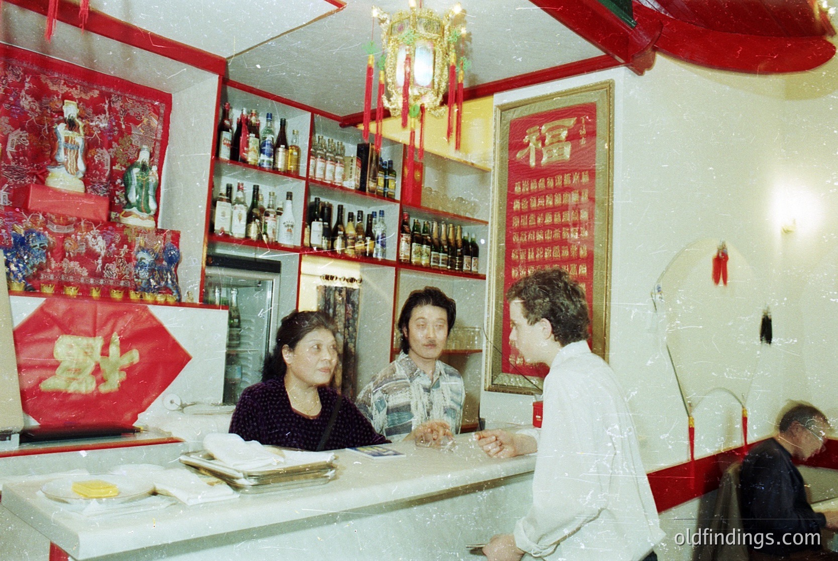 Vintage interior of a Chinese restaurant or teahouse, featuring red lacquered walls, shelves stocked with bottled drinks, and framed calligraphy. Three patrons seated at a counter, one man in a light-colored shirt and two women in dark attire. Decorative red lanterns and a hanging golden ornament add cultural detail. Likely