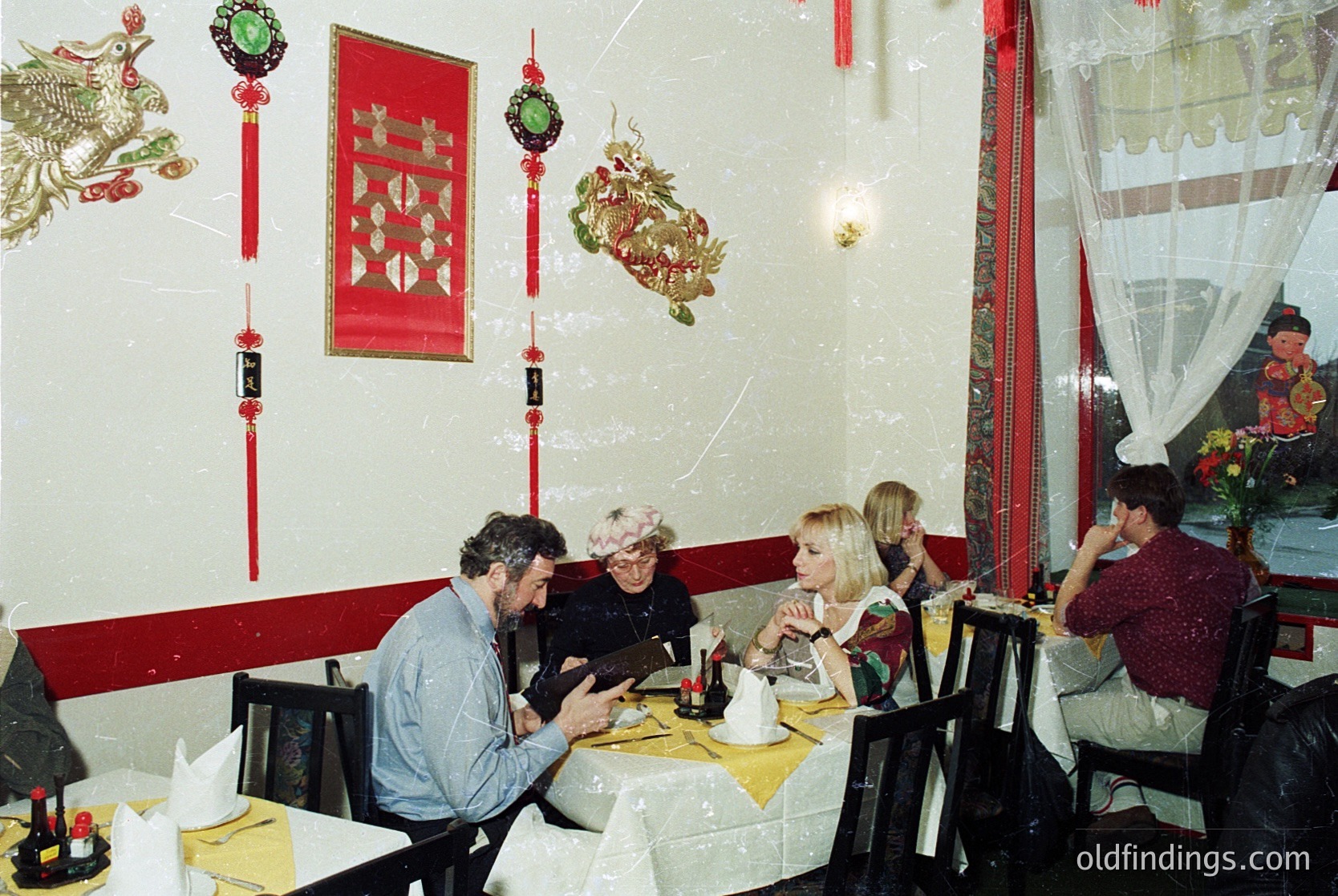 Vintage Asian-themed restaurant interior with red-and-gold decor, featuring dragon and shield wall hangings. Four adults seated at round tables with yellow tablecloths, engaging in conversation. Mid-20th century style with floral curtains and classic tableware. Likely --- *Note: Exact location indeterminable, but decor suggests East/Southeast Asian influence.*