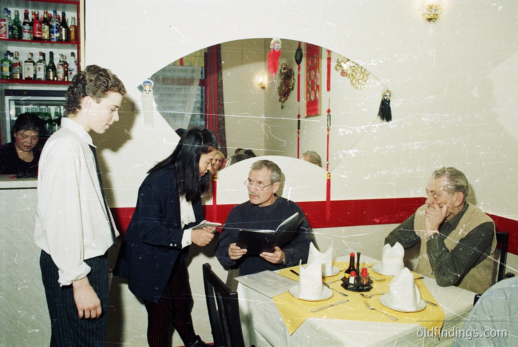 Vintage indoor scene: four adults in a dimly lit restaurant, likely 1970s–1980s. A waiter in white shirt and black apron hands a menu to a woman in a dark blazer. Seated patrons—one man in a sweater vest, another in a jacket—engage in conversation. Shelves behind display bottles, and a red-and-white archway frames a mirror with decorative items. Warm, intimate dining atmosphere.