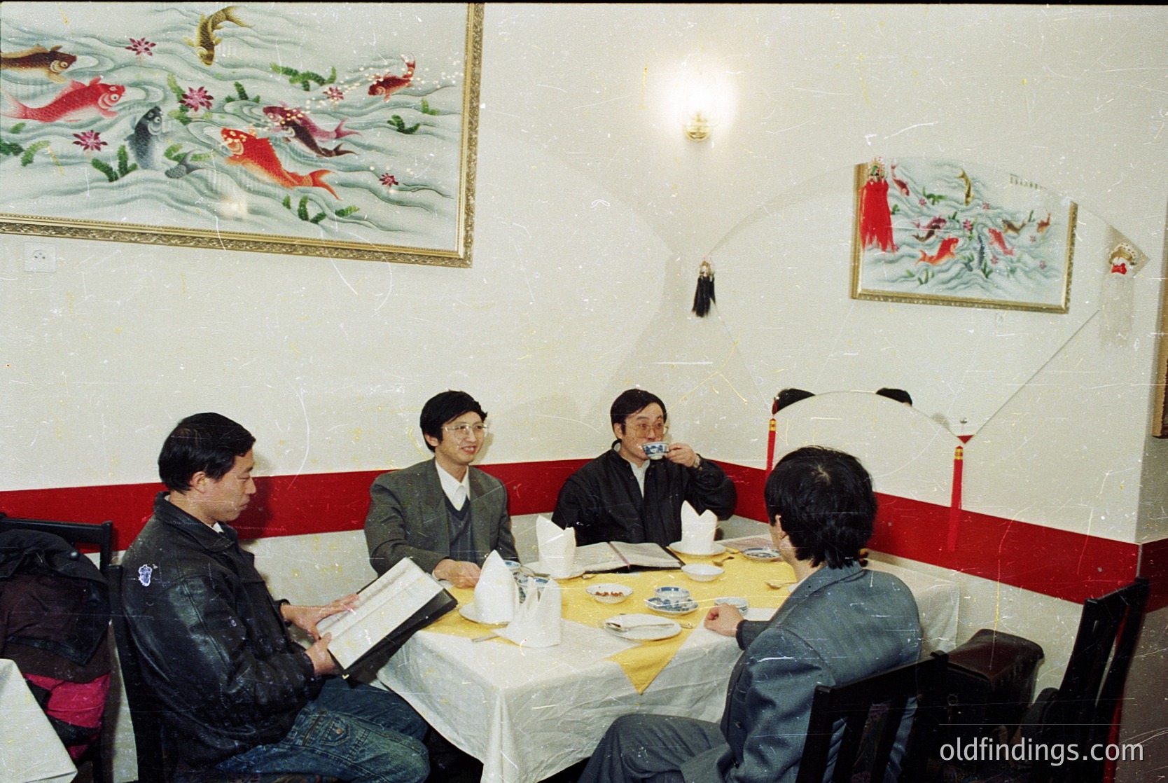 Four men in formal attire gather around a table in a dimly lit, traditional-style restaurant. Decorative koi fish paintings adorn the cream walls with red accents. Yellow tablecloths and white plates suggest a mid-meal setting. Style hints at East Asian dining culture, likely 1980s–1990s.