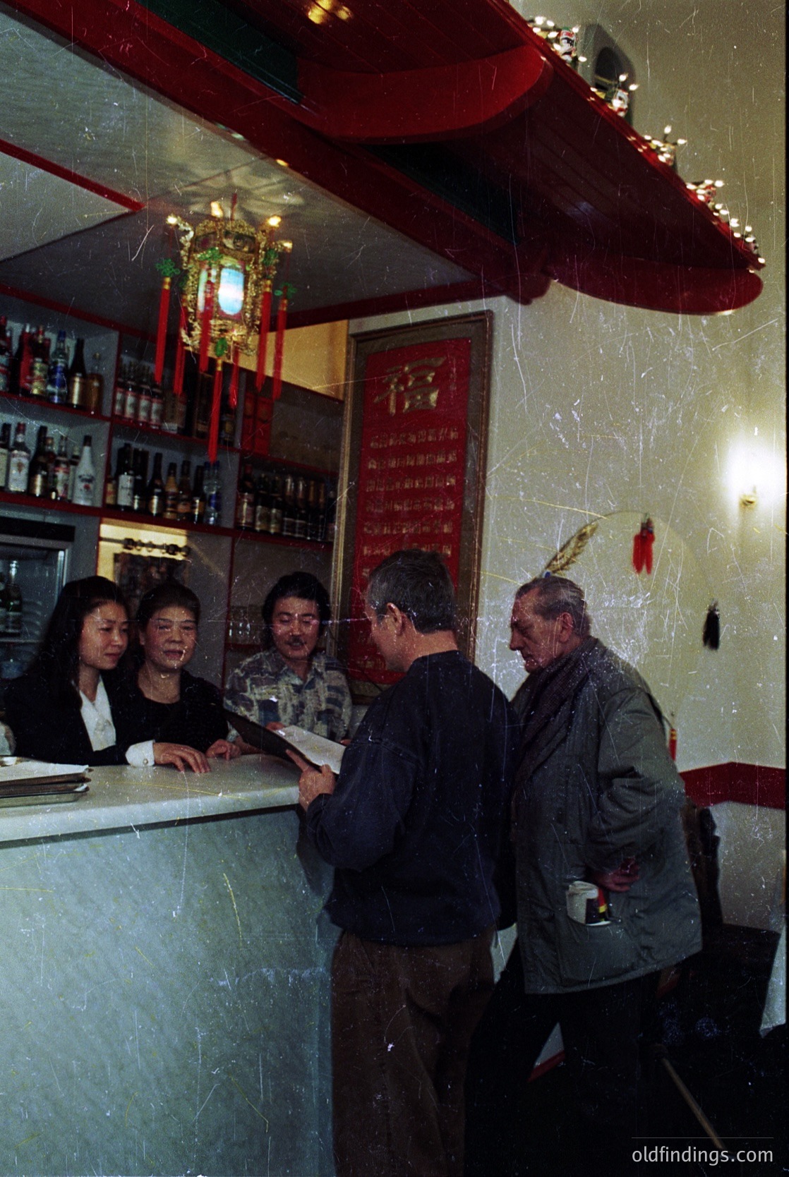 Vintage bar scene with five patrons at a counter adorned with red vinyl wallpaper and Chinese characters. Bottles line shelves behind the bar, and a red lantern hangs above. Men in jackets and women in formal attire suggest a mid-20th century setting, likely 1950s–1960s.