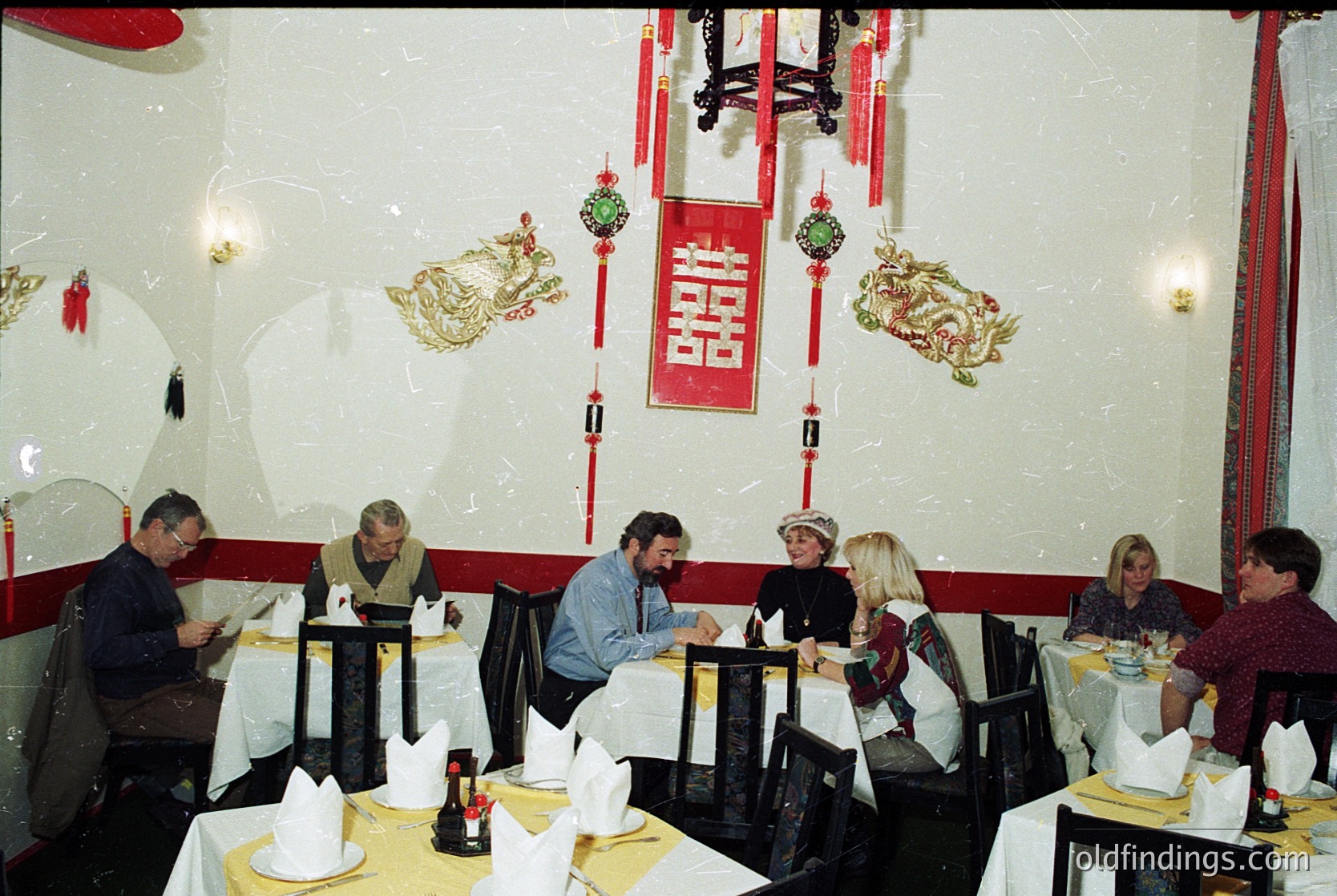 Mid-century Asian-themed restaurant interior with red-and-gold dragon motifs and calligraphy. Round tables, black chairs, and white tablecloths set for dining. Group of seven adults seated, engaged in conversation. Decor includes red lanterns, hanging ornaments, and a central red banner with Chinese characters. Likely 1970s–1980s Western setting.