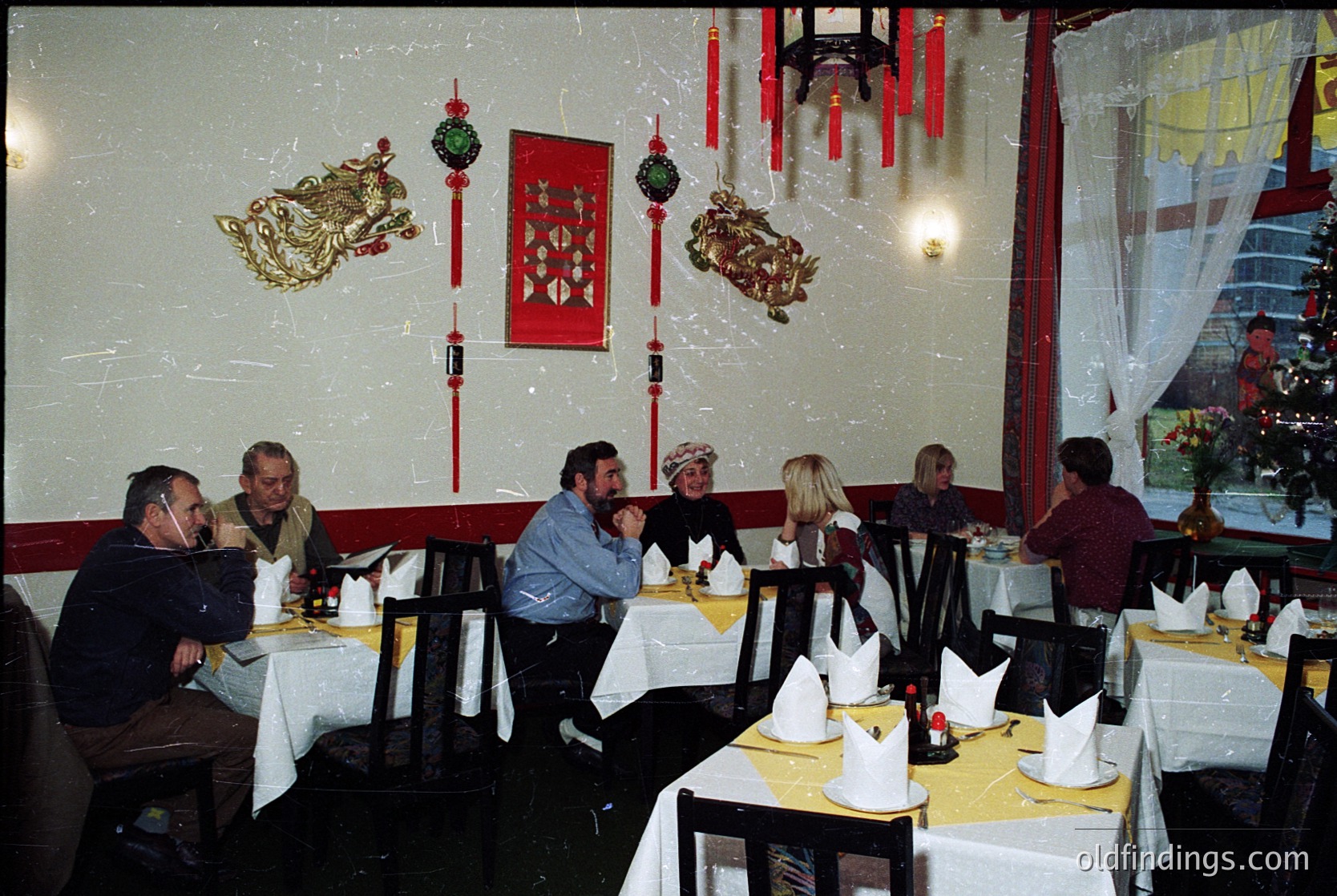 Vintage restaurant interior with Asian-inspired decor: round tables draped in white tablecloths, red lanterns, and gold dragon motifs. Eight seated patrons in 1970s-80s attire (blazers, vests, headscarves) dining under warm lighting. Reflective glass wall reveals outdoor greenery. éVibes