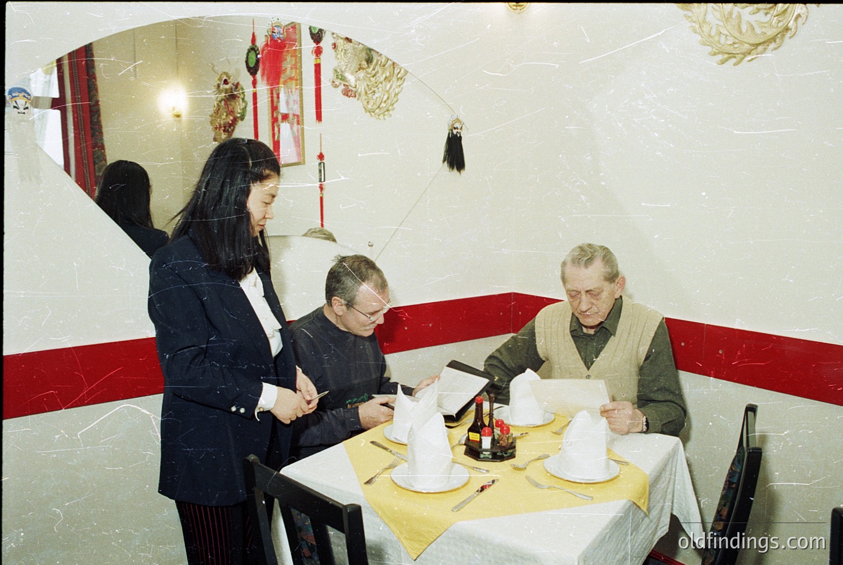 Vintage indoor dining scene with three adults seated at a round table in a restaurant. The woman stands, holding a menu, while two men review documents. Decor includes red-and-white striped wall trim, a large circular mirror, and red Chinese New Year decorations. Mid-century tableware and formal attire suggest a 1960s–1970s setting.