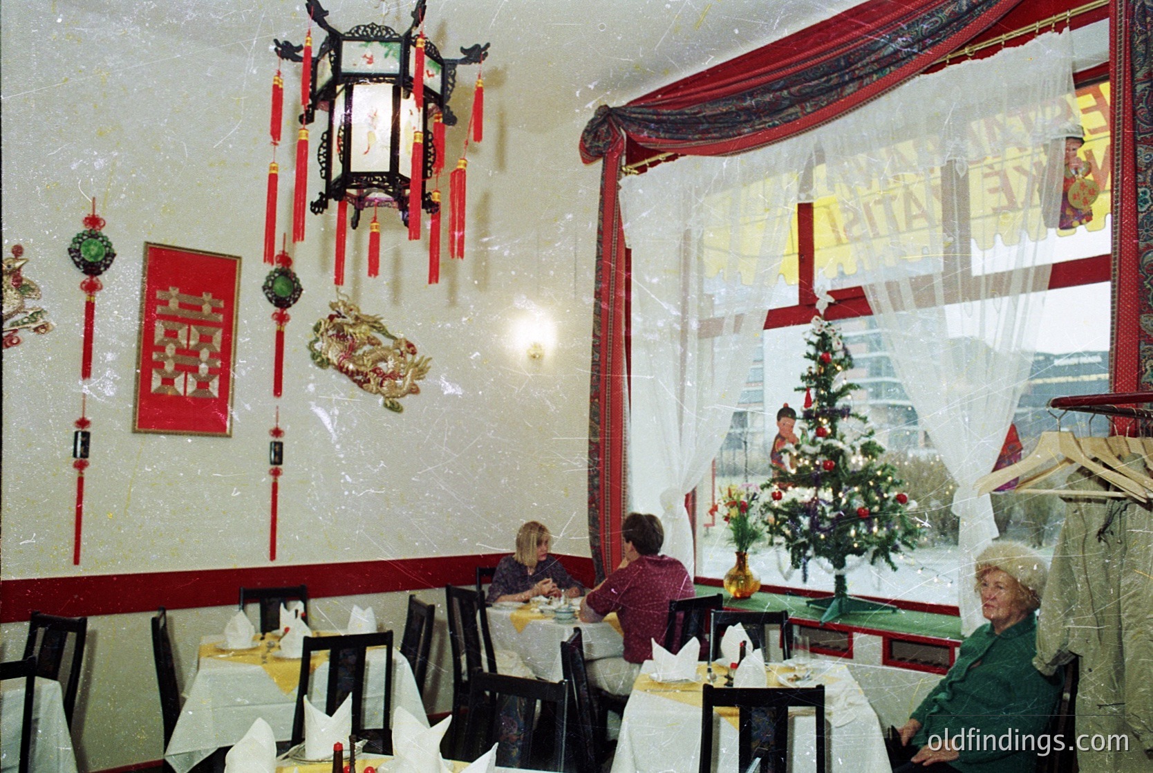 Vintage Chinese-themed restaurant interior with red lanterns, calligraphy, and festive Christmas tree. Frosted windows reveal snowy urban backdrop. Mid-century dining setup with white tablecloths and red upholstery.