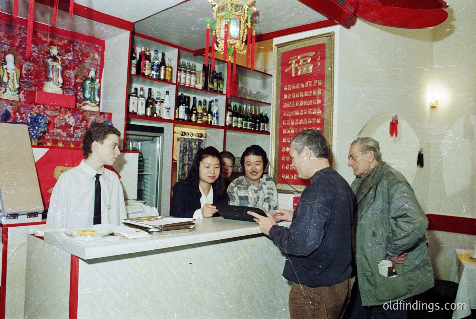 Vintage interior of a Chinese restaurant or teahouse, featuring a counter with shelves stocked with bottled beverages and framed red banners with Chinese calligraphy. Five individuals—two men in casual winter wear, two women in business attire, and one male server in a white shirt—engage in conversation. Decor includes red wall panels and traditional red lanterns. Likely