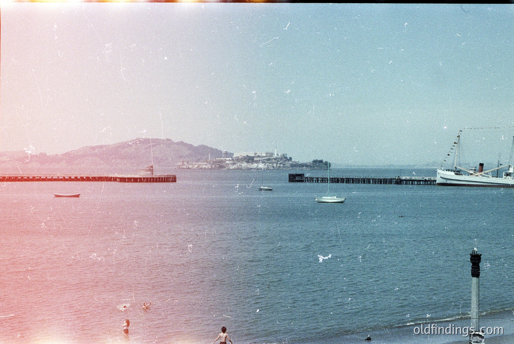 Vintage seaside pier extending into calm waters, flanked by small boats. Distant urban coastline with low-rise buildings and a large ship anchored offshore. Sunlight casts warm tones, suggesting late afternoon. Likely mid-20th century coastal scene.