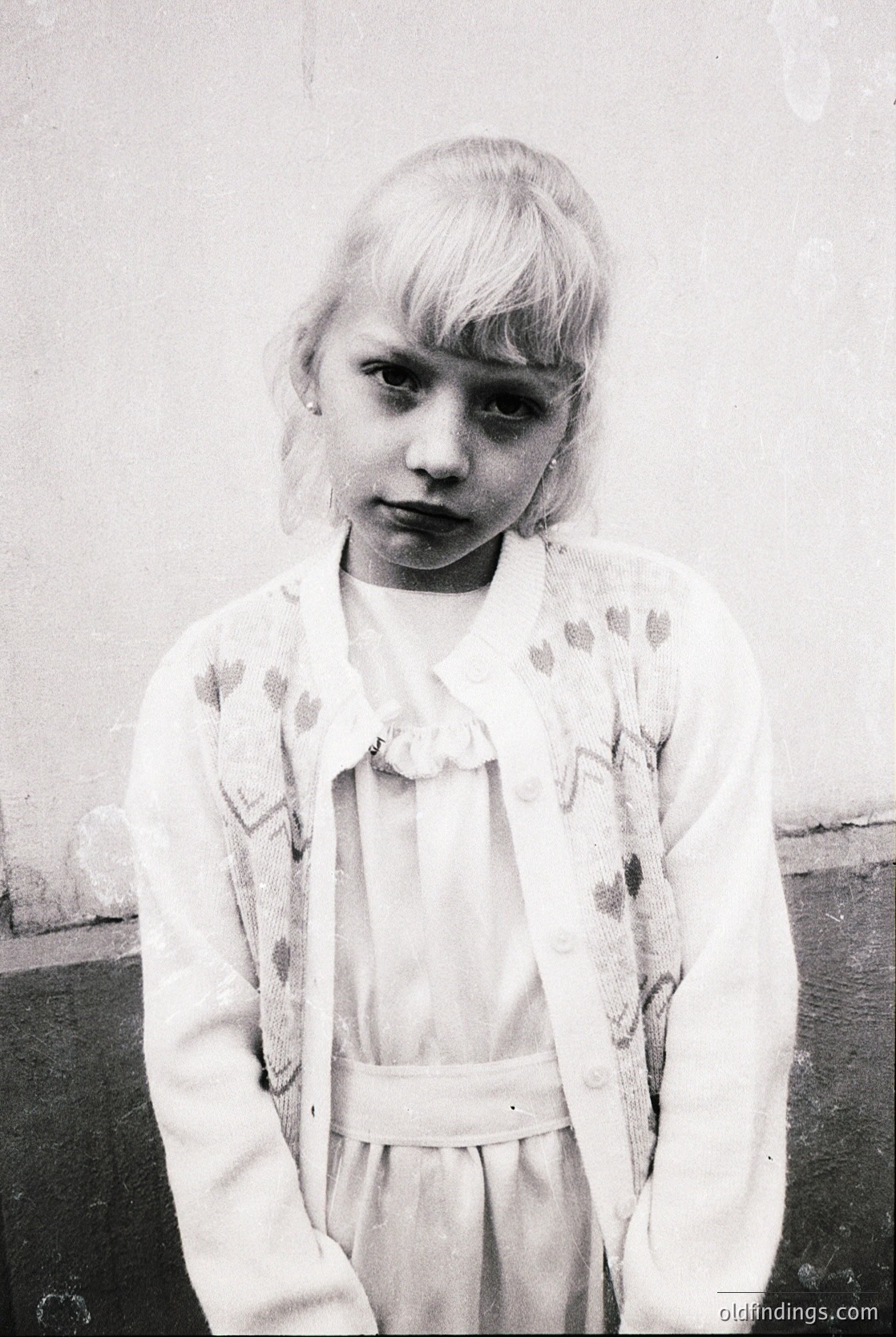 Portrait of a young girl in mid-20th century clothing—white floral cardigan over a dress with a belted waist. Plain concrete wall background suggests urban or institutional setting. Style hints at 1950s–1960s Western or Eastern European fashion.
