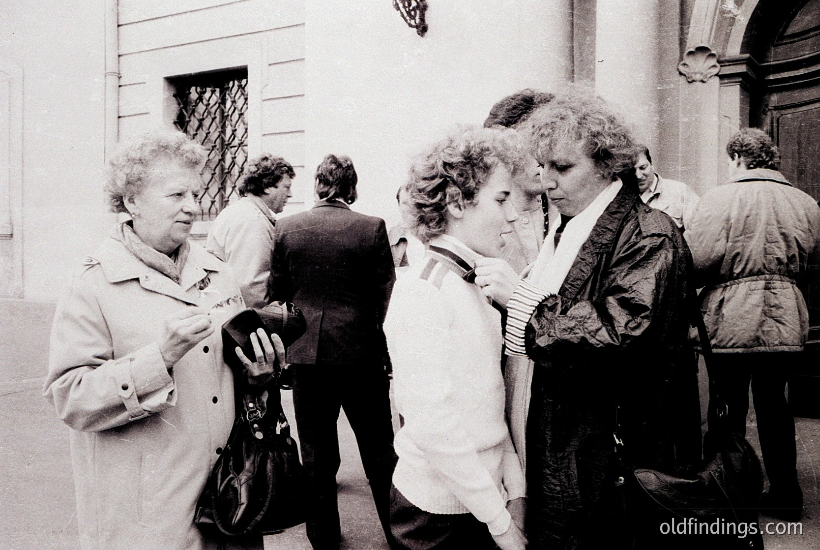 Black-and-white candid of three women in mid-conversation outside a building with classical architectural details. Women wear 1970s-era coats, scarves, and handbags. Crowd of blurred figures in formal attire suggests an event or gathering.
