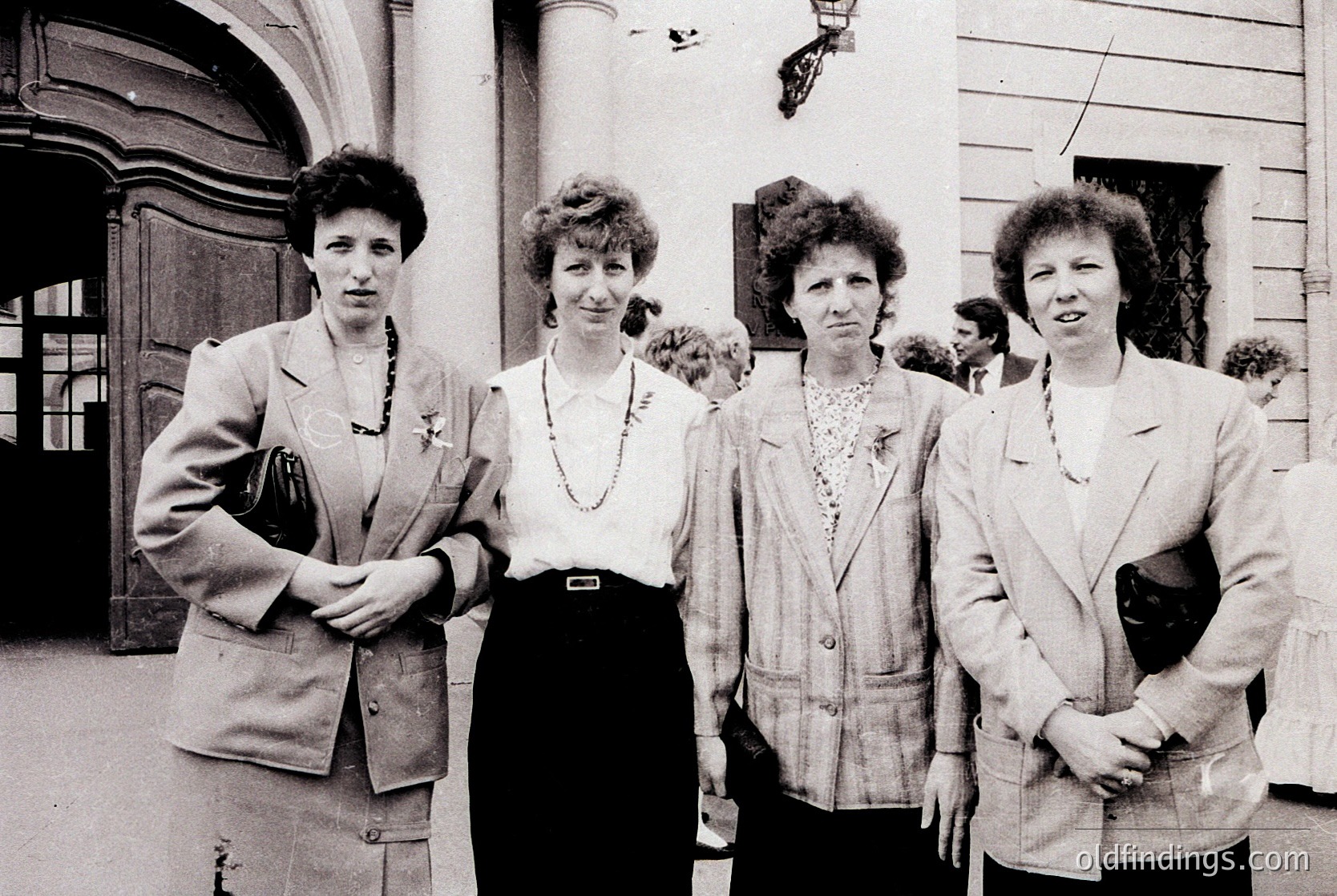 Four women in 1970s-era blazers and blouses pose outdoors near a grand, arched doorway. Formal attire suggests a professional or official gathering. Architectural details hint at institutional or governmental setting.