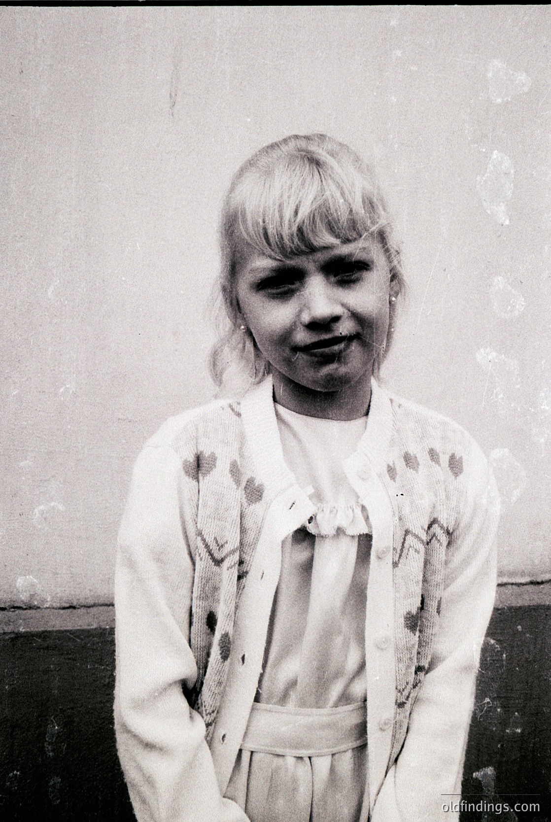 Young girl in 1960s-70s fashion, posing against a plain wall. She wears a floral-patterned blouse with a belted cardigan, short bangs, and a slight smile. Candid, timeless portrait style.