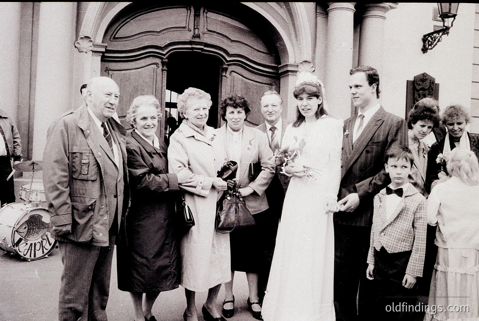Black-and-white wedding photo outside a grand, arched entrance with classical columns. A bride in a simple white gown holds flowers, surrounded by family in 1960s-70s attire—men in suits, women in dresses and headscarves. Formal, candid moment captures generational presence.