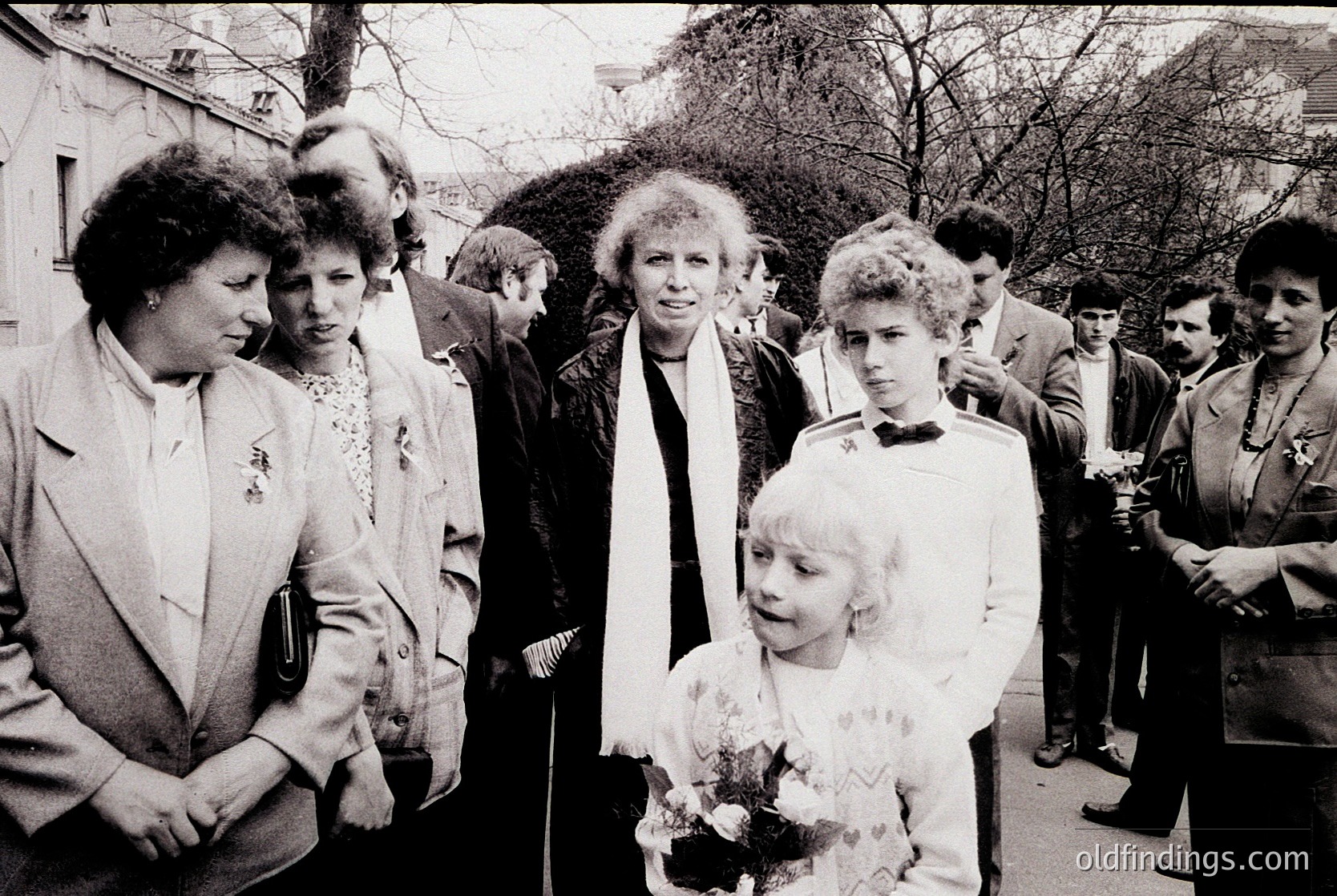 Black-and-white street scene featuring a group of adults and children in formal attire, likely 1970s–1980s. Central figure in a white coat holds a bouquet; others wear blazers and scarves. Urban park or campus setting with bare trees and stone buildings in background.