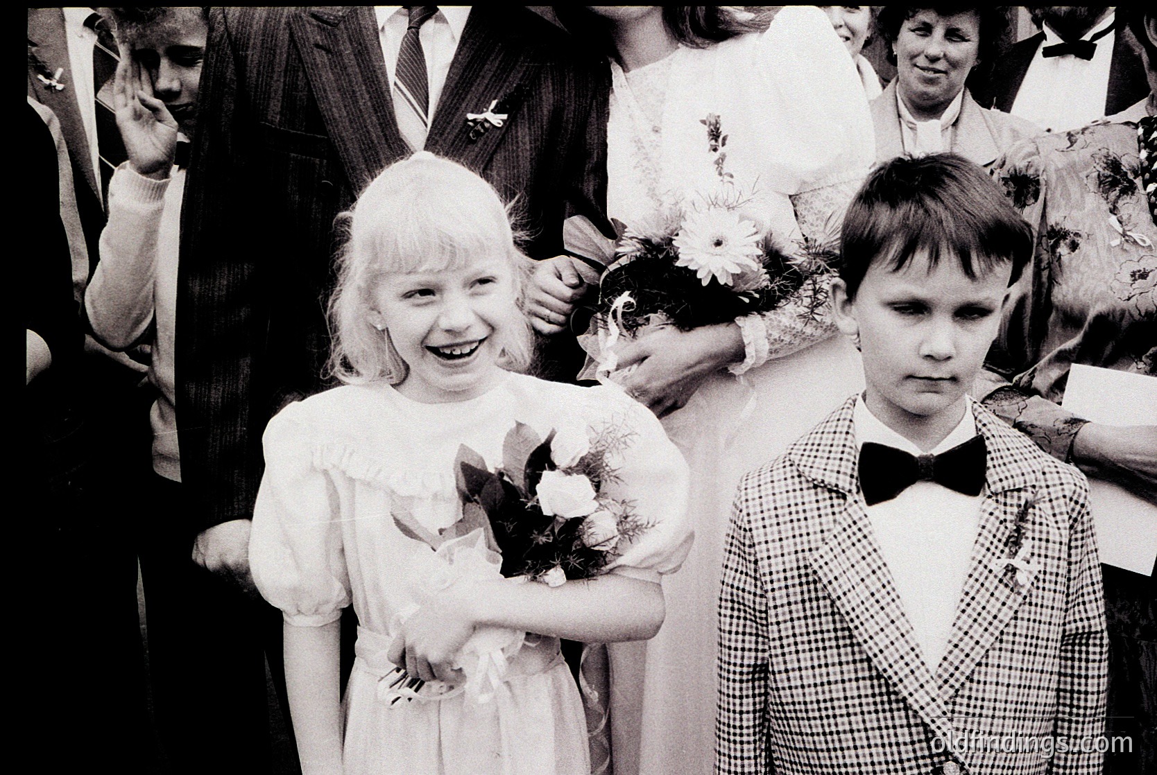 Young girl and boy in formal attire, likely 1950s–1970s Western/European wedding. Girl wears a simple white dress with bouquet; boy in checkered suit with bow tie. Candid, joyful expressions capture innocence and tradition.