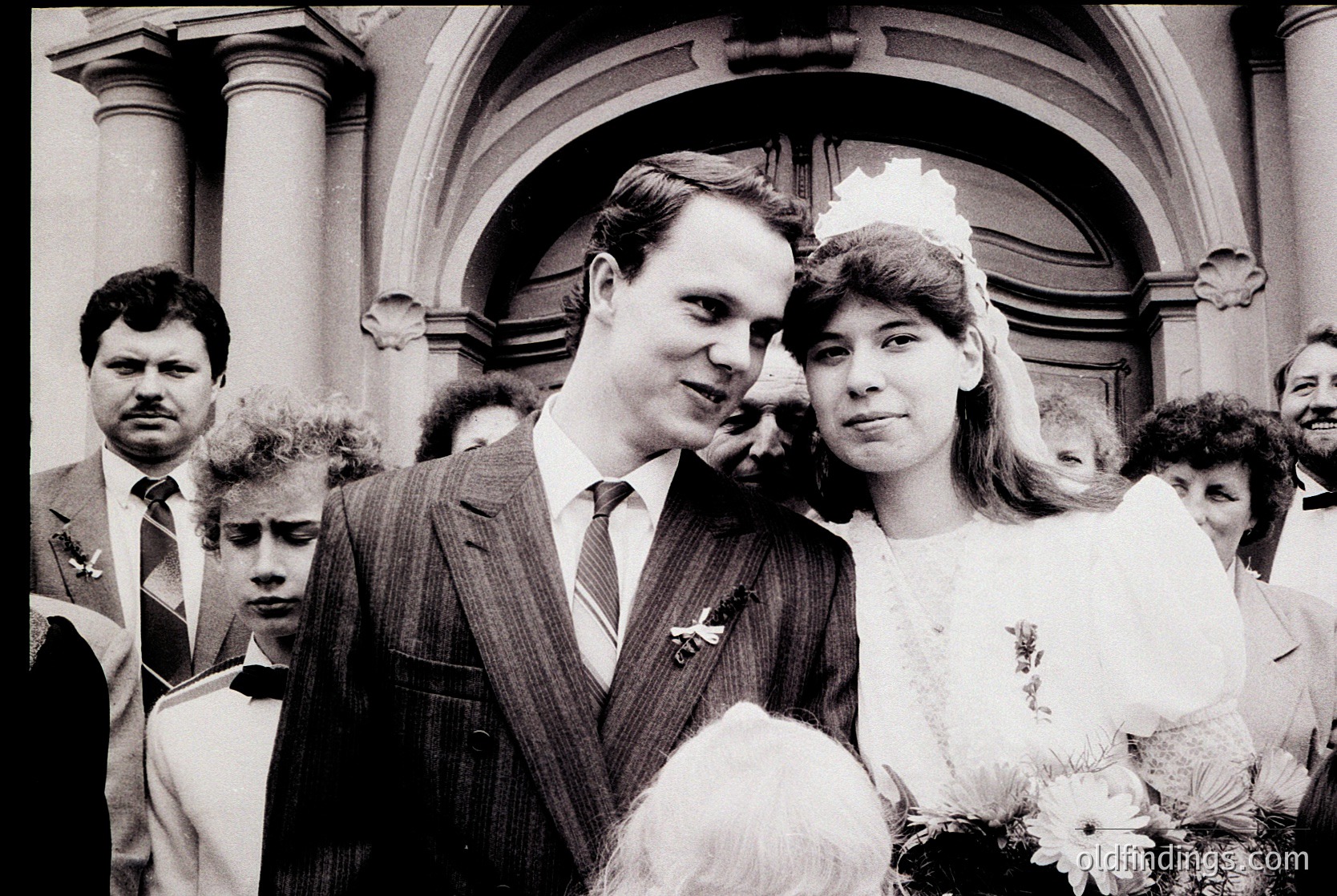 Classic black-and-white wedding photo featuring a bride in a simple white gown with veil and bouquet, posing with groom in a suit outside a grand, arched entrance. Crowd in formal attire suggests 1970s–1980s European ceremony. Architectural details hint at historic or institutional venue.