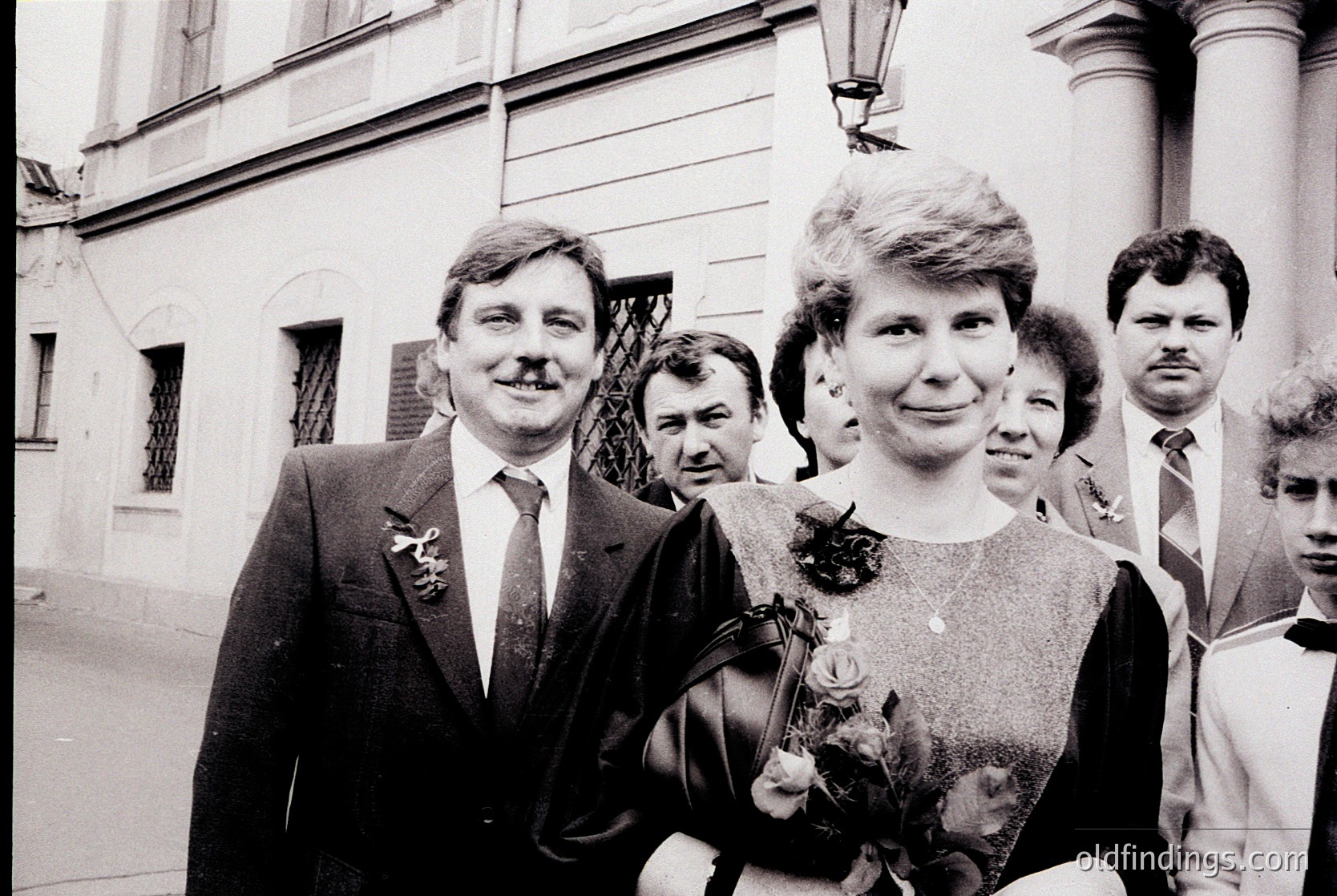 Black-and-white portrait of a formal gathering in Eastern Europe, likely 1970s–1980s. Central couple in formal attire: man in suit with lapel pin, woman in dress holding floral bouquet. Architectural backdrop features classical columns and stone facade. Candid expressions suggest a celebratory or official event.