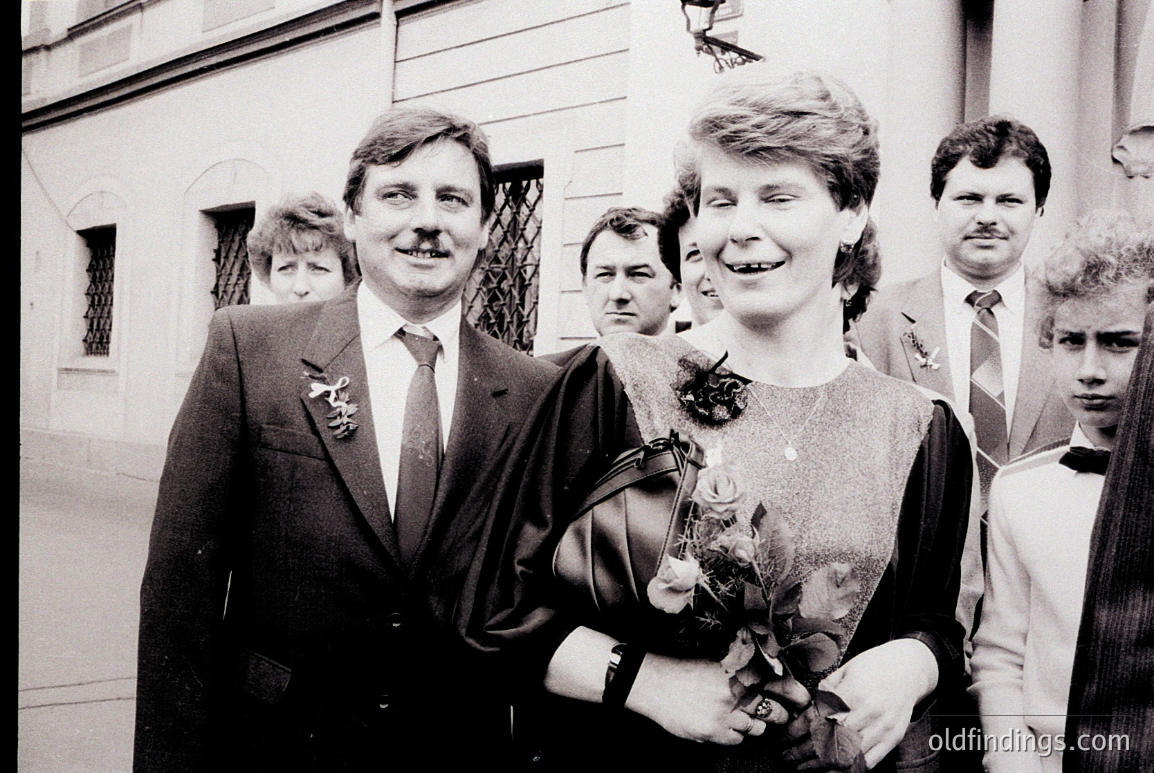 Couple in formal attire at what appears to be a wedding, holding floral bouquets. Man in suit with boutonnière, woman in knee-length dress with corsage. Urban setting with classical architecture in background. Likely 1970s–1980s Western Europe.