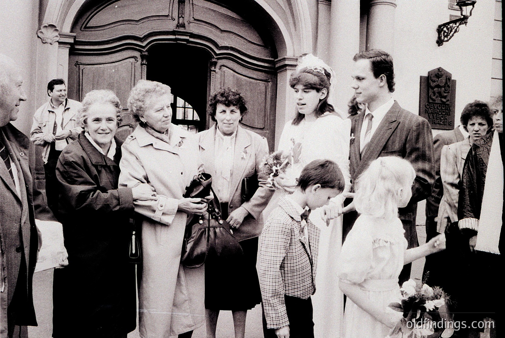 Black-and-white photo of a wedding procession outside a grand, arched doorway, likely Eastern European . Six adults in formal attire (suits, dresses) and two children in school uniforms, holding bouquets. Architectural details include ornate wrought-iron railings and a classical facade.