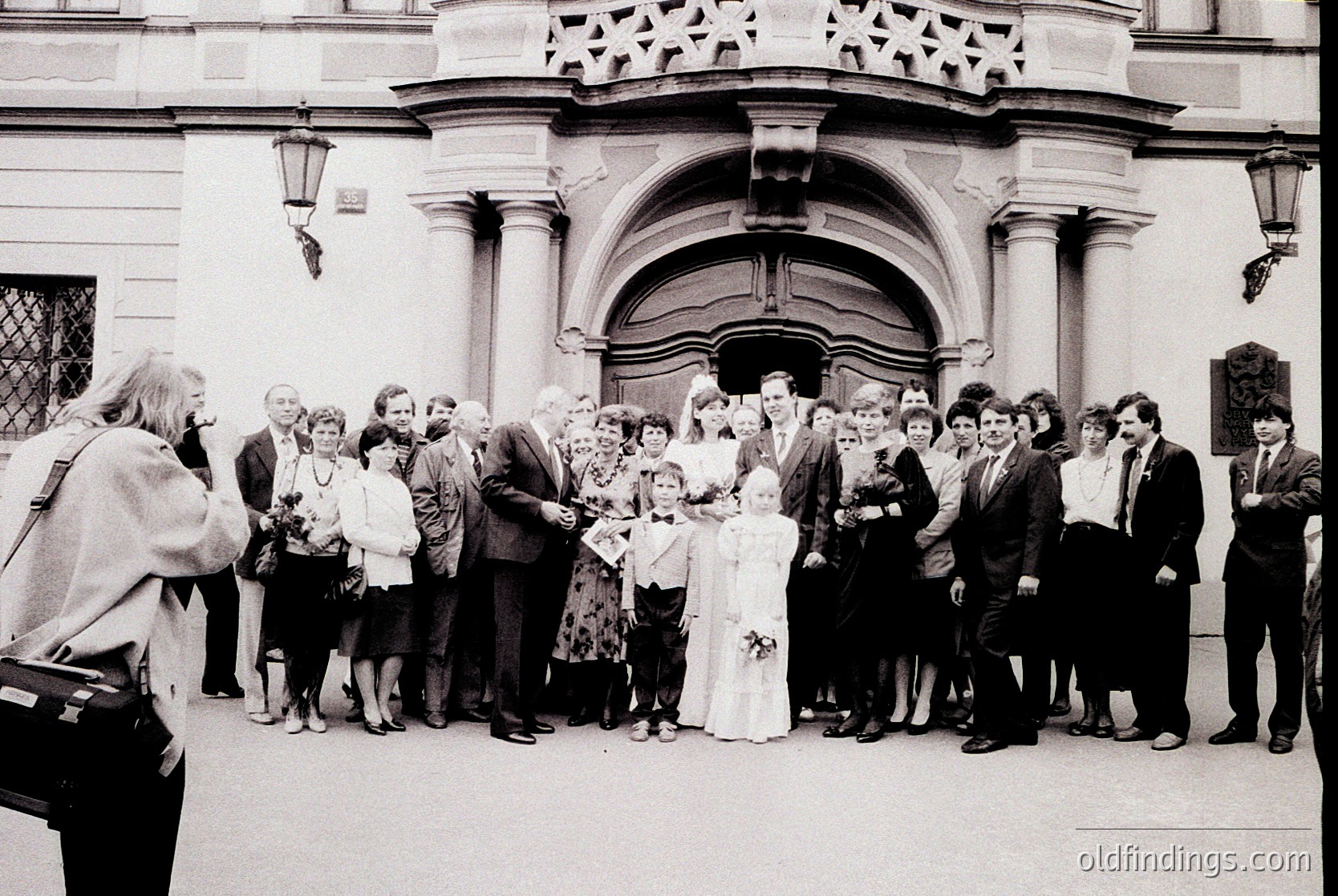 Neoclassical building entrance with arched doorway and decorative stonework hosts a formal gathering. A bride in white stands center, surrounded by guests in 1970s-era attire—men in suits, women in dresses and headscarves. One guest holds a camera, capturing the moment. Crowded urban setting suggests a wedding or celebratory event.