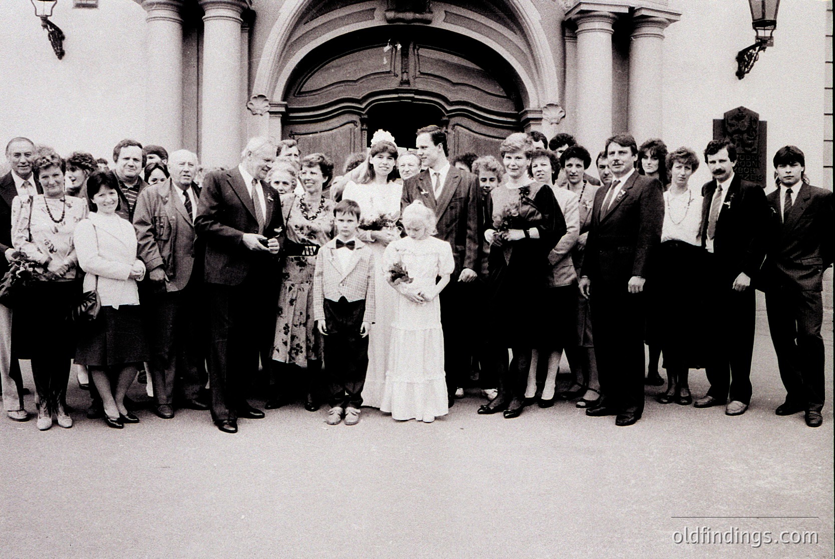 Group wedding photo in grand neoclassical building entrance, likely Eastern Bloc era (1970s–1980s). Bride in simple white gown, groom in suit, surrounded by 16 guests in formal attire. Floral bouquets held by attendees. Architectural details: arched doorway, columns, and ornate street lamps.
